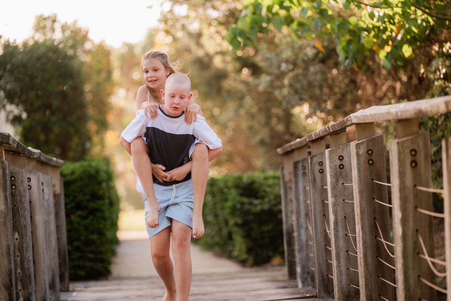 Candid photo of two children playing on a wooden bridge outdoors, with one giving the other a piggyback ride. Tweed Coast photography.