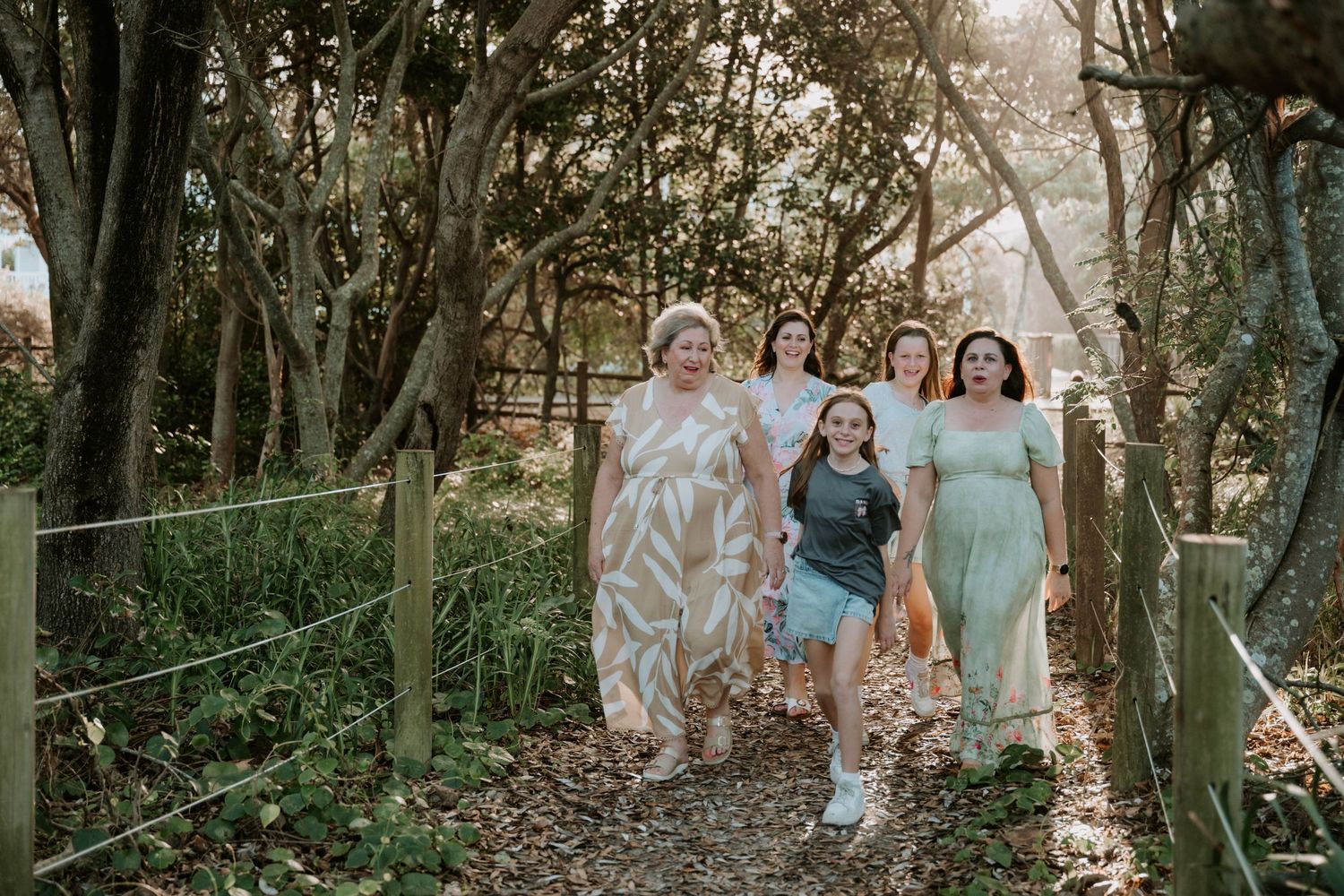 Five women walking together on a sunlit forest path surrounded by lush green trees and wooden fencing.