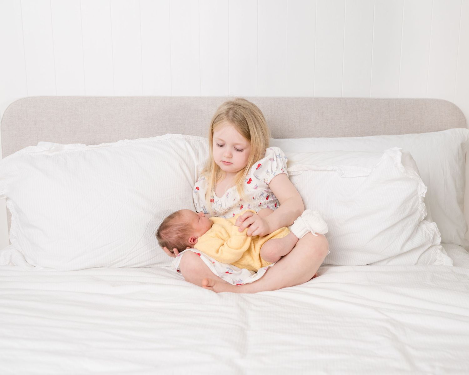 A young blonde girl sits on a white bed, gently cradling a newborn baby wrapped in a yellow swaddle.