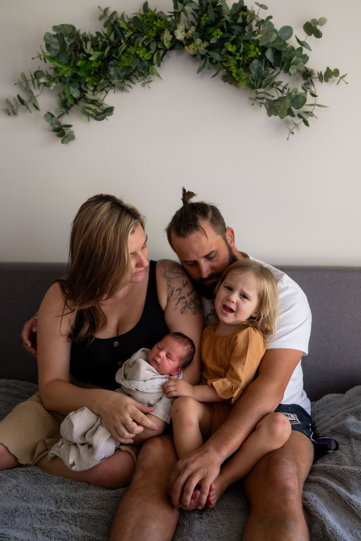 Family of four sitting on bed with newborn baby, toddler girl, mom and tattooed dad under eucalyptus arch.