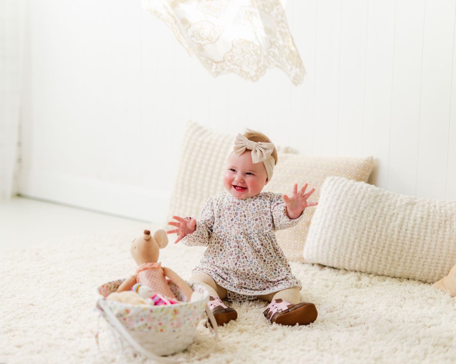 Happy baby girl in floral outfit sitting on white fluffy rug, laughing and reaching up toward floating lace decoration.