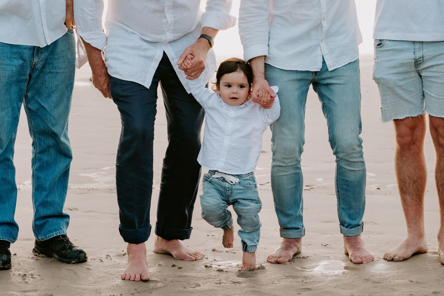 Toddler learning to walk on beach held by two adults in jeans, barefoot on wet sand.