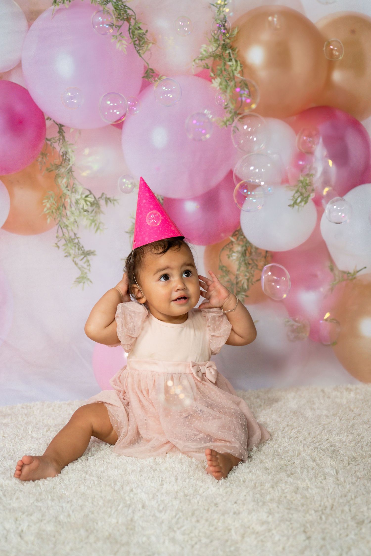 Baby in pink dress sits on white rug with pink and gold balloon arch and greenery decorations above.