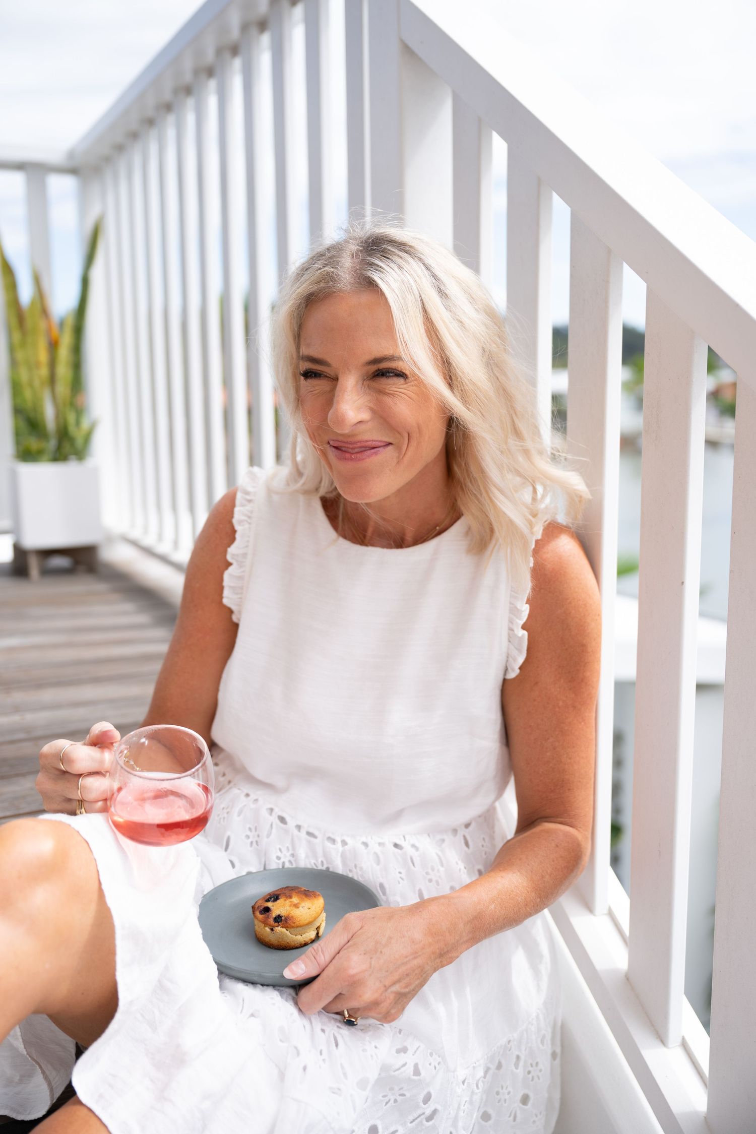 Smiling blonde woman in white dress holding rose wine and small pastry on balcony with white railing.