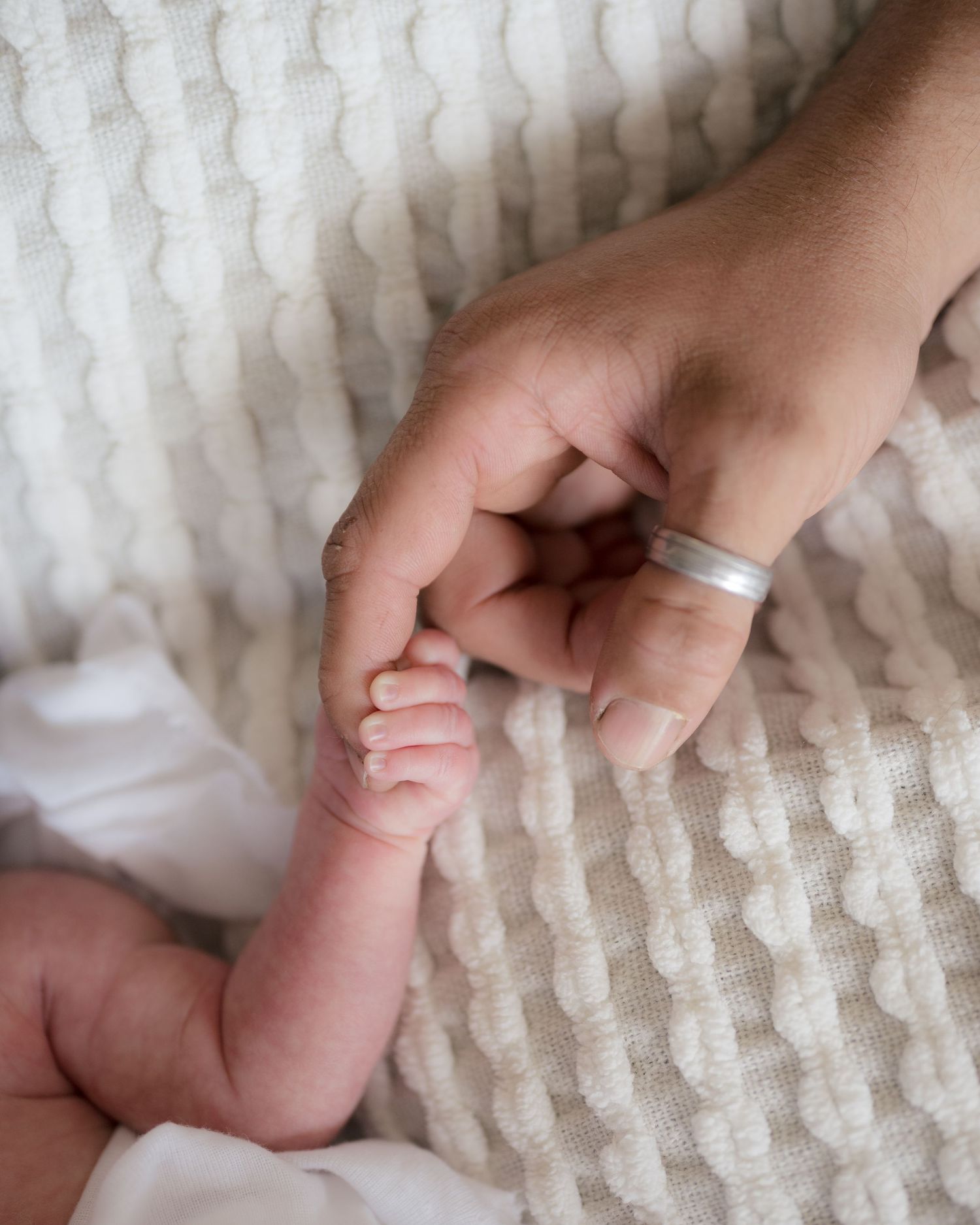 A newborn baby grasping an adult's finger wearing a silver ring on a soft white textured blanket.