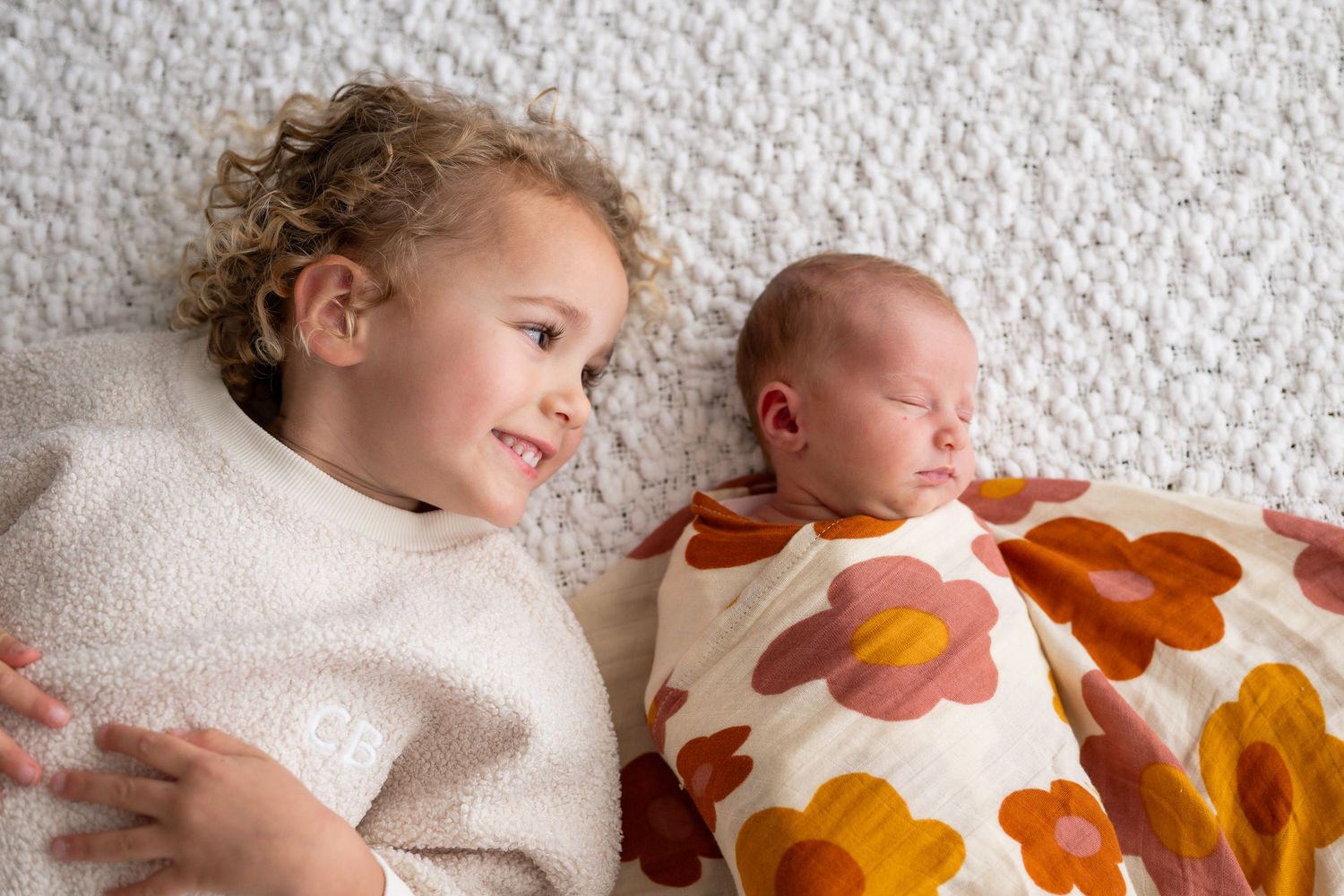 Older toddler with curly hair lying beside sleeping newborn wrapped in floral swaddle blanket.