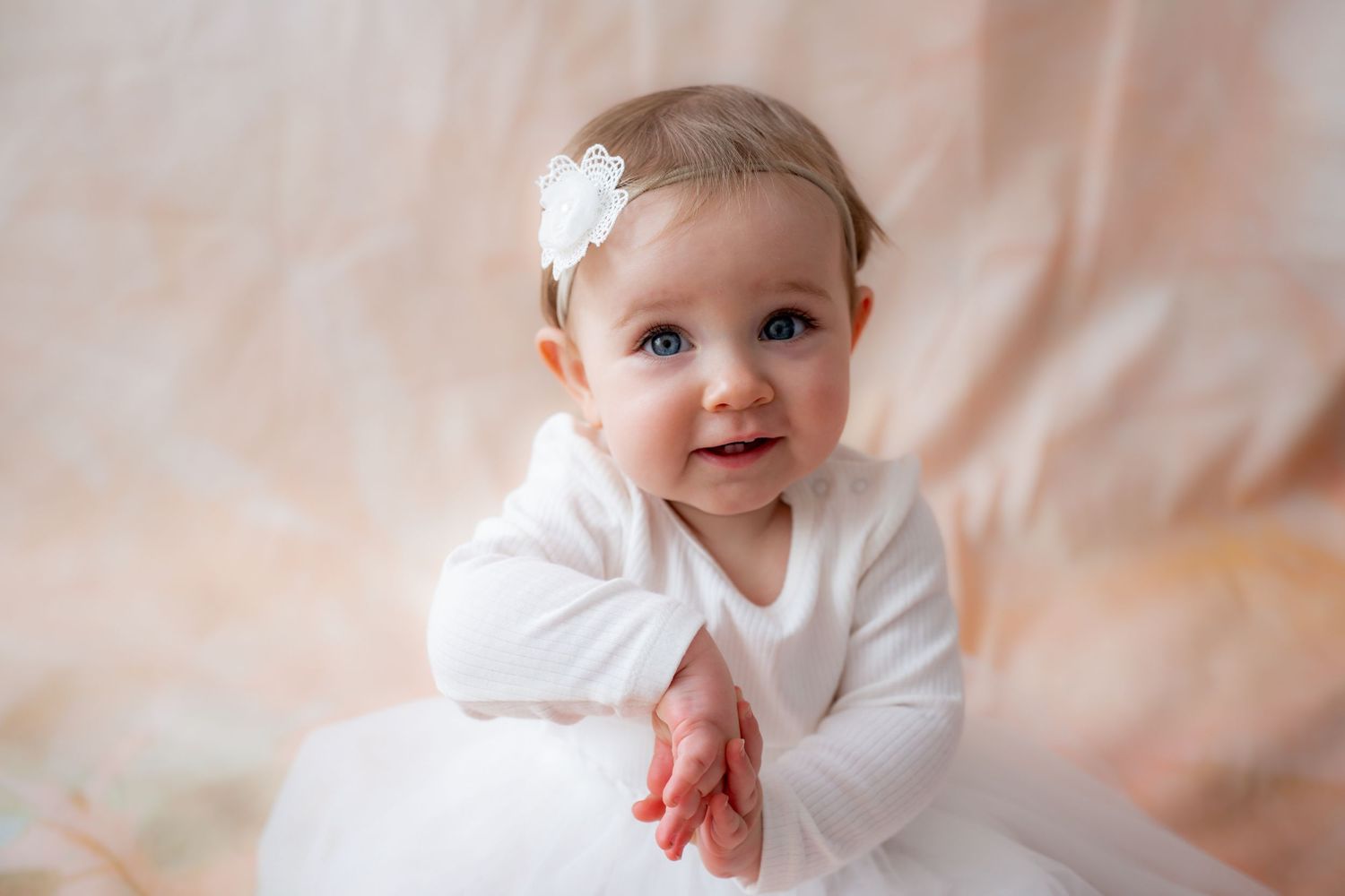 Adorable baby girl in white dress and floral headband smiling during a soft pink studio portrait session.