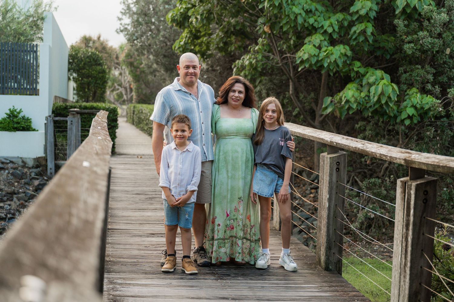 Family of four smiling on a wooden boardwalk surrounded by lush greenery on a sunny day.