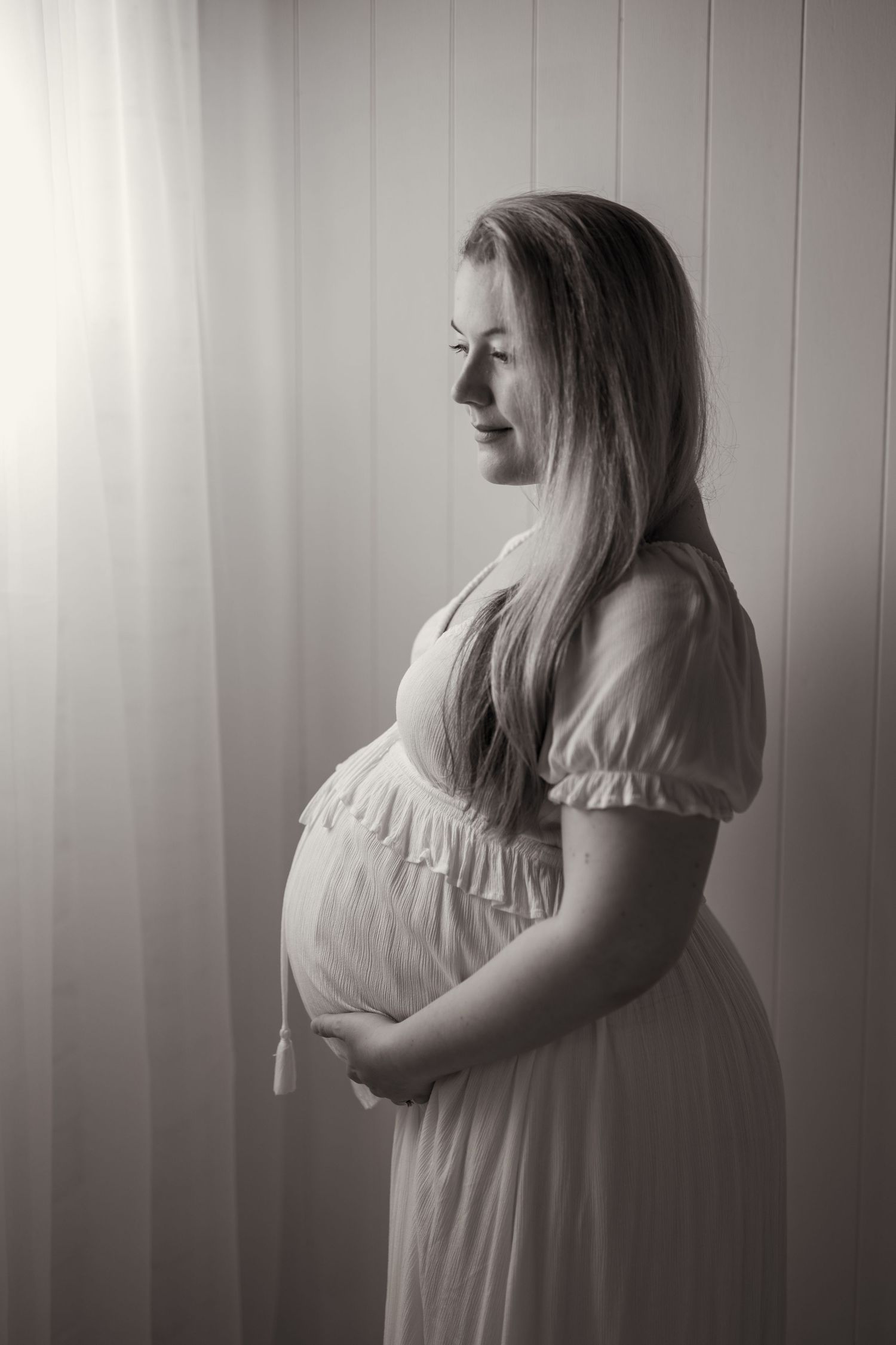 Black and white portrait of a pregnant woman in a flowing dress, cradling her baby bump by a sunlit window.