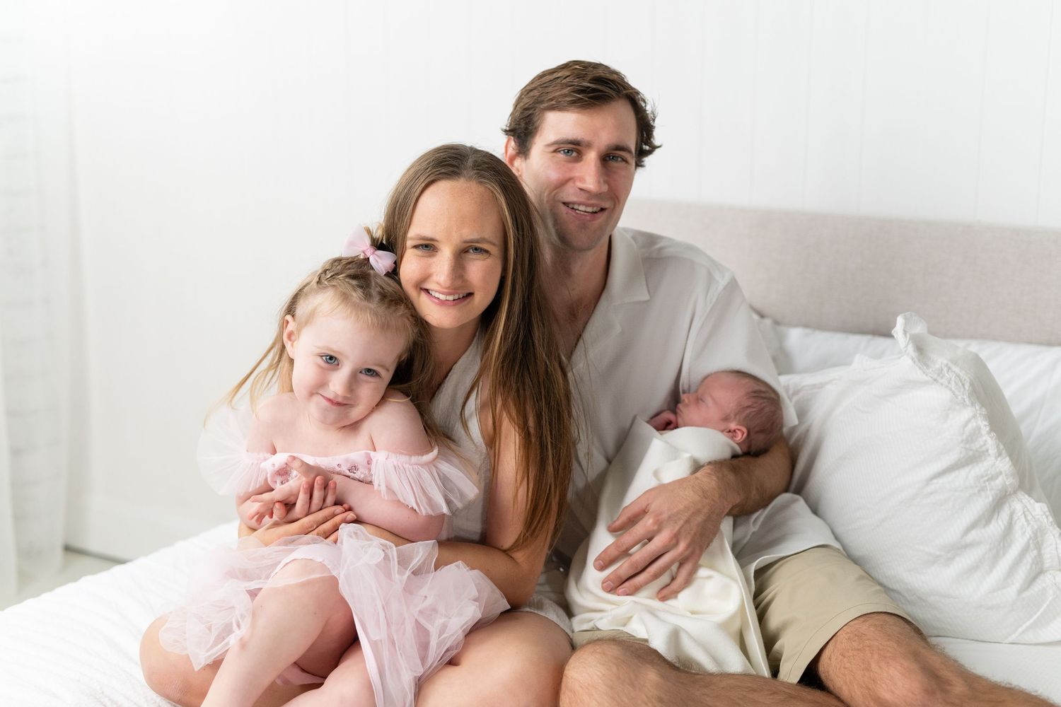Family of four posing on a white bed, parents smiling with toddler girl in pink dress and newborn baby.