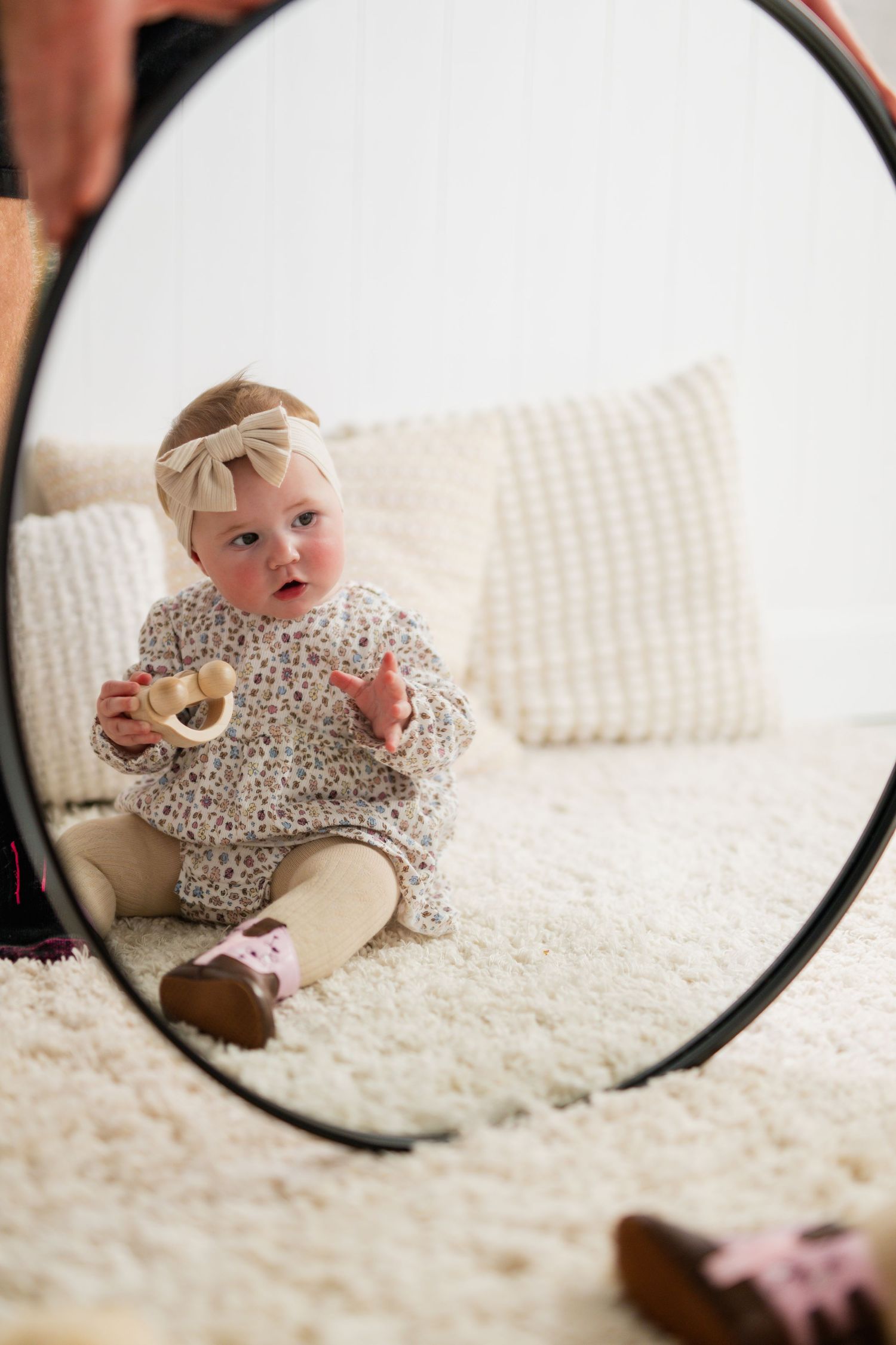 Baby girl in floral dress and bow headband sits on fluffy rug, holding wooden toy, reflected in round mirror.
