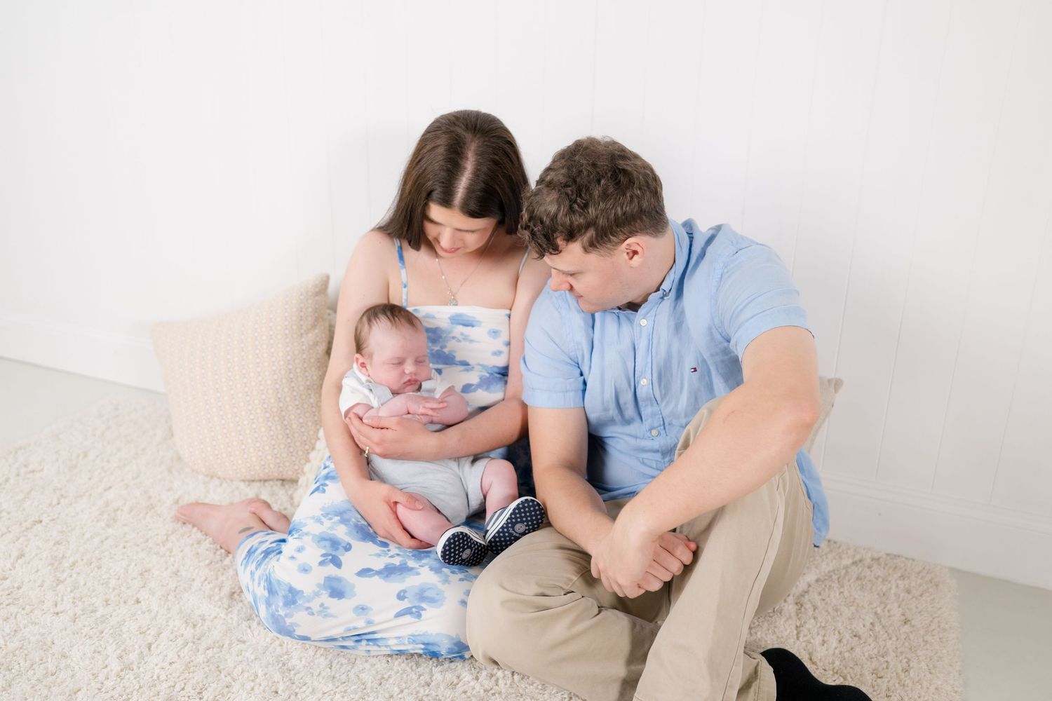 New parents sitting on a white rug, lovingly gazing at their newborn baby wrapped in a blue floral swaddle.