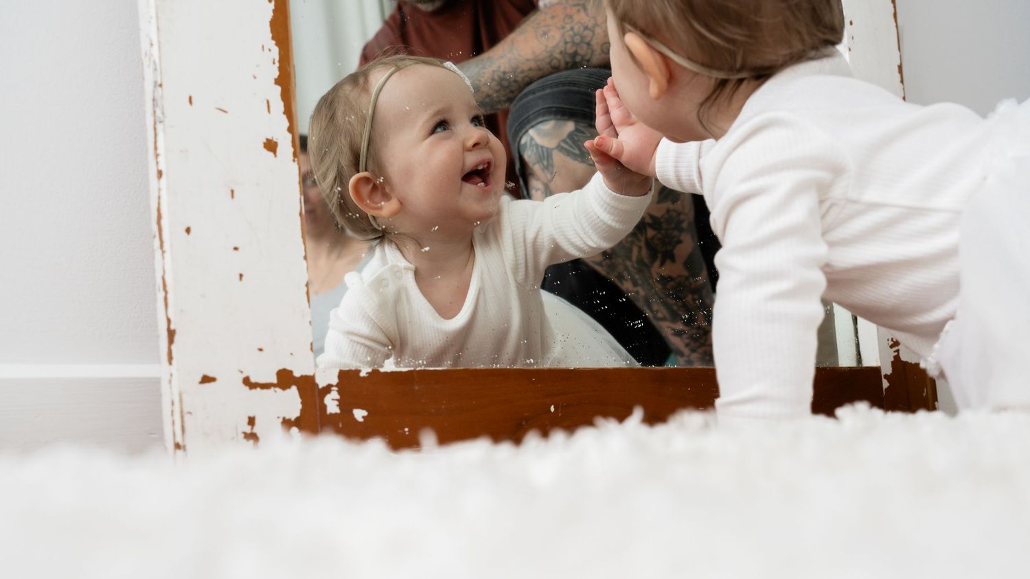A happy toddler in white outfit touches mirror reflection, smiling joyfully on fluffy white carpet.