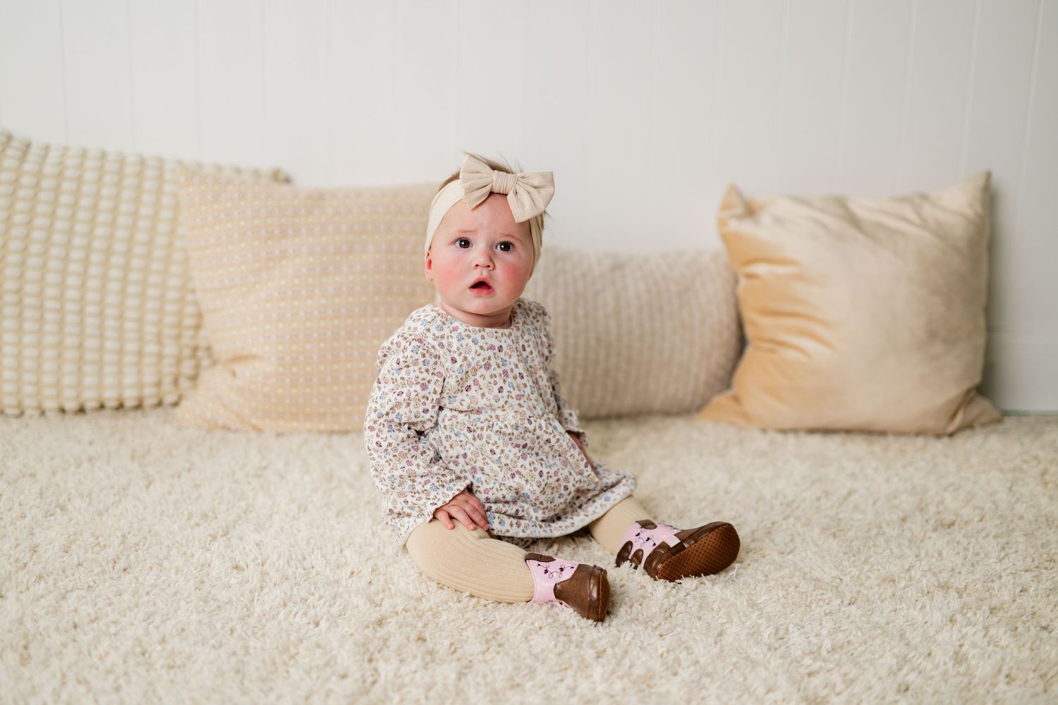 Adorable baby girl in floral outfit and bow headband sitting on a cream textured surface with pillows.