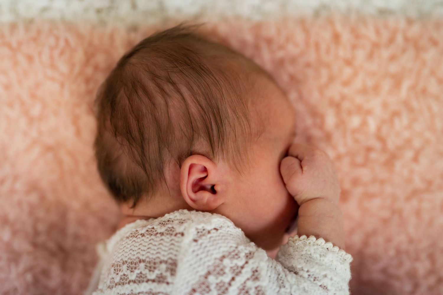 Newborn baby sleeping peacefully on a pink blanket, wearing a white lace outfit, with tiny hand near face.