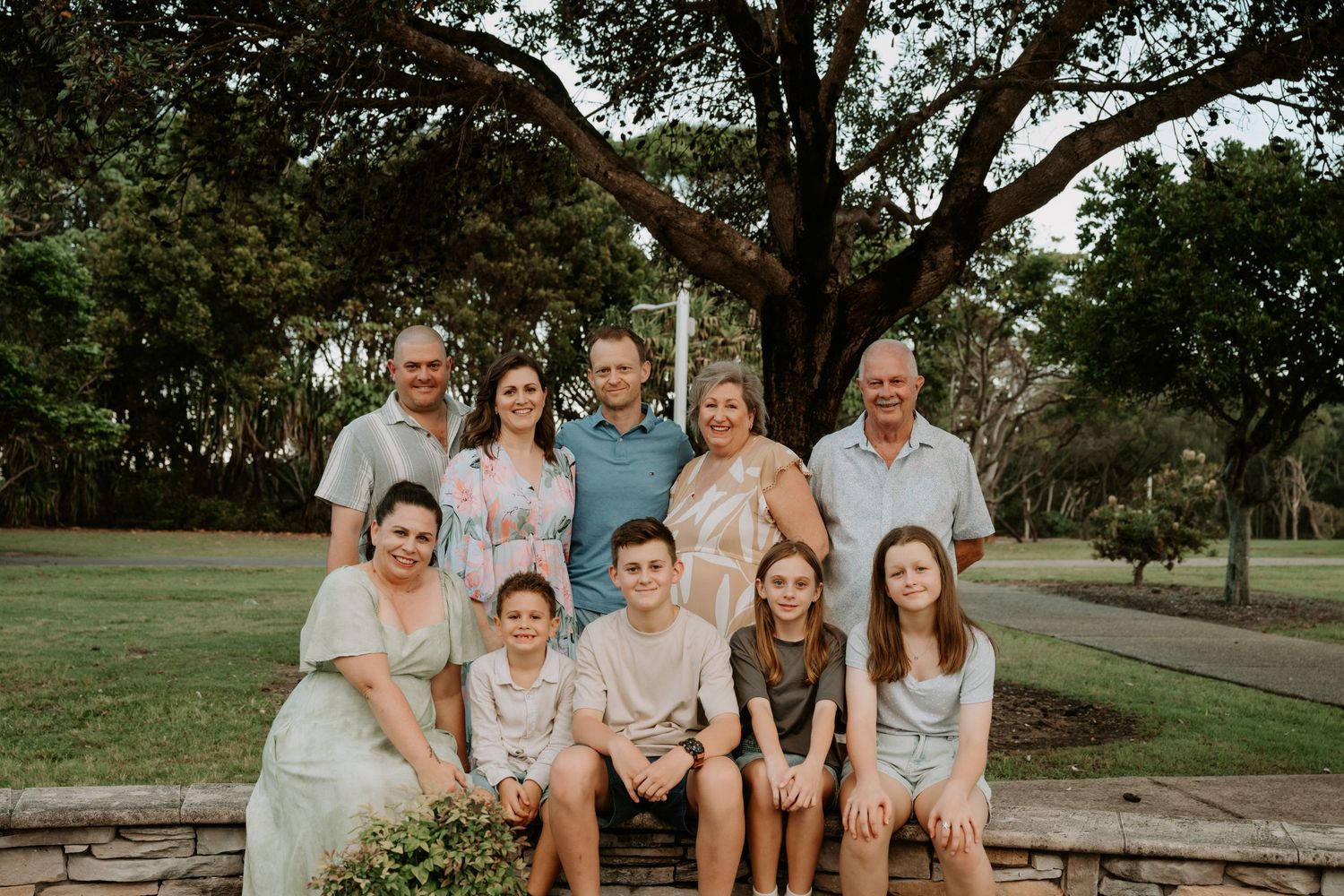 Multi-generational family group portrait outdoors, smiling together near a large tree on green grass.