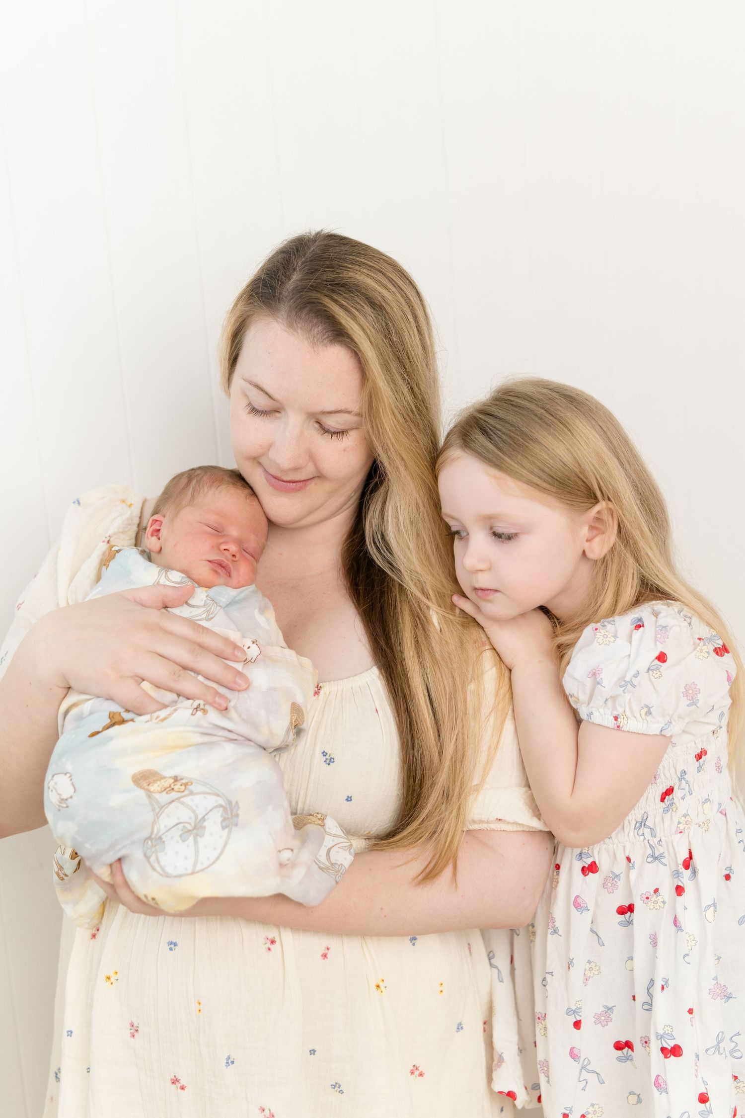 A mother in a yellow dress holds her swaddled newborn while her young blonde daughter gazes lovingly at the baby.