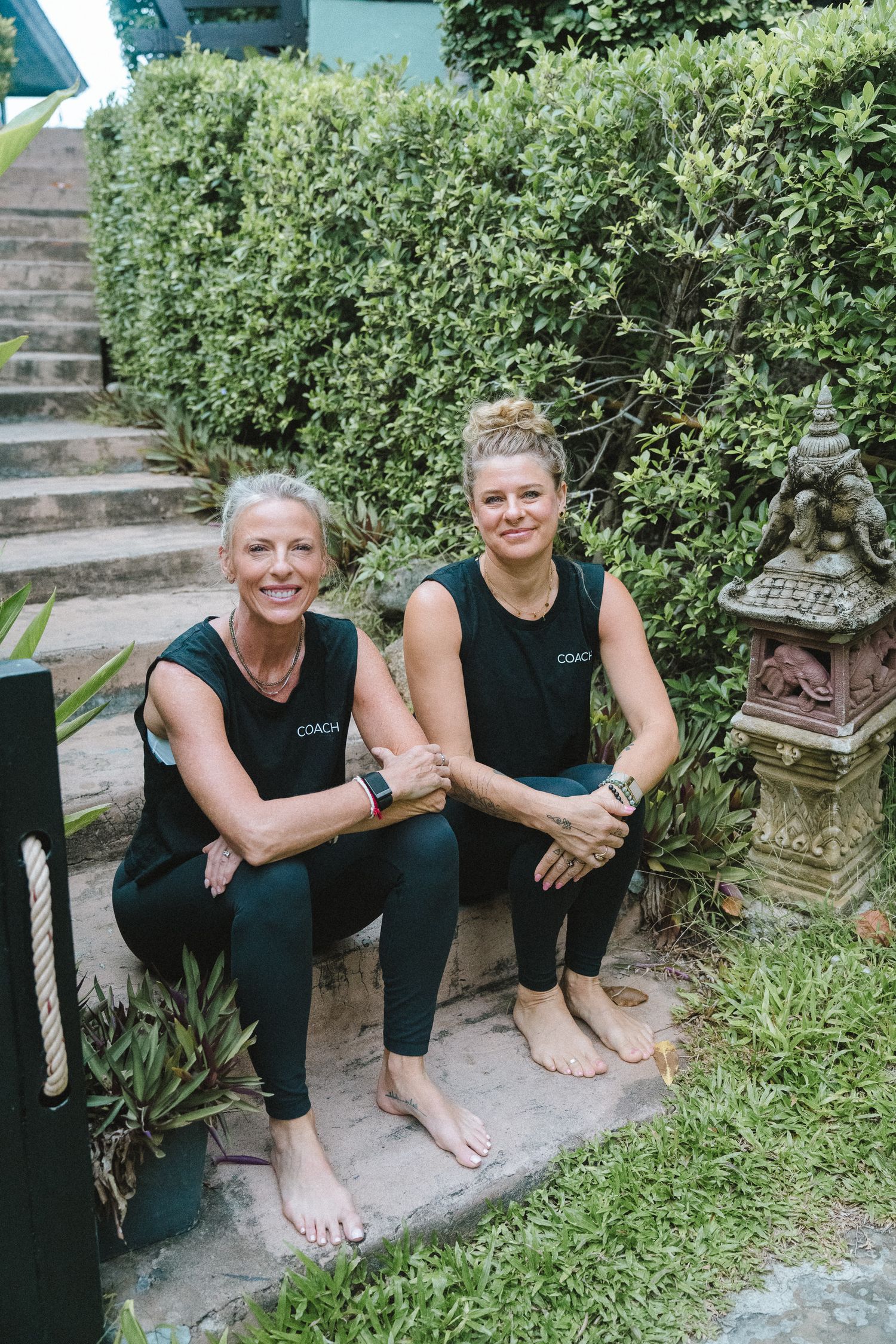 Two smiling women in matching dark athletic wear sitting barefoot on garden steps surrounded by tropical greenery.