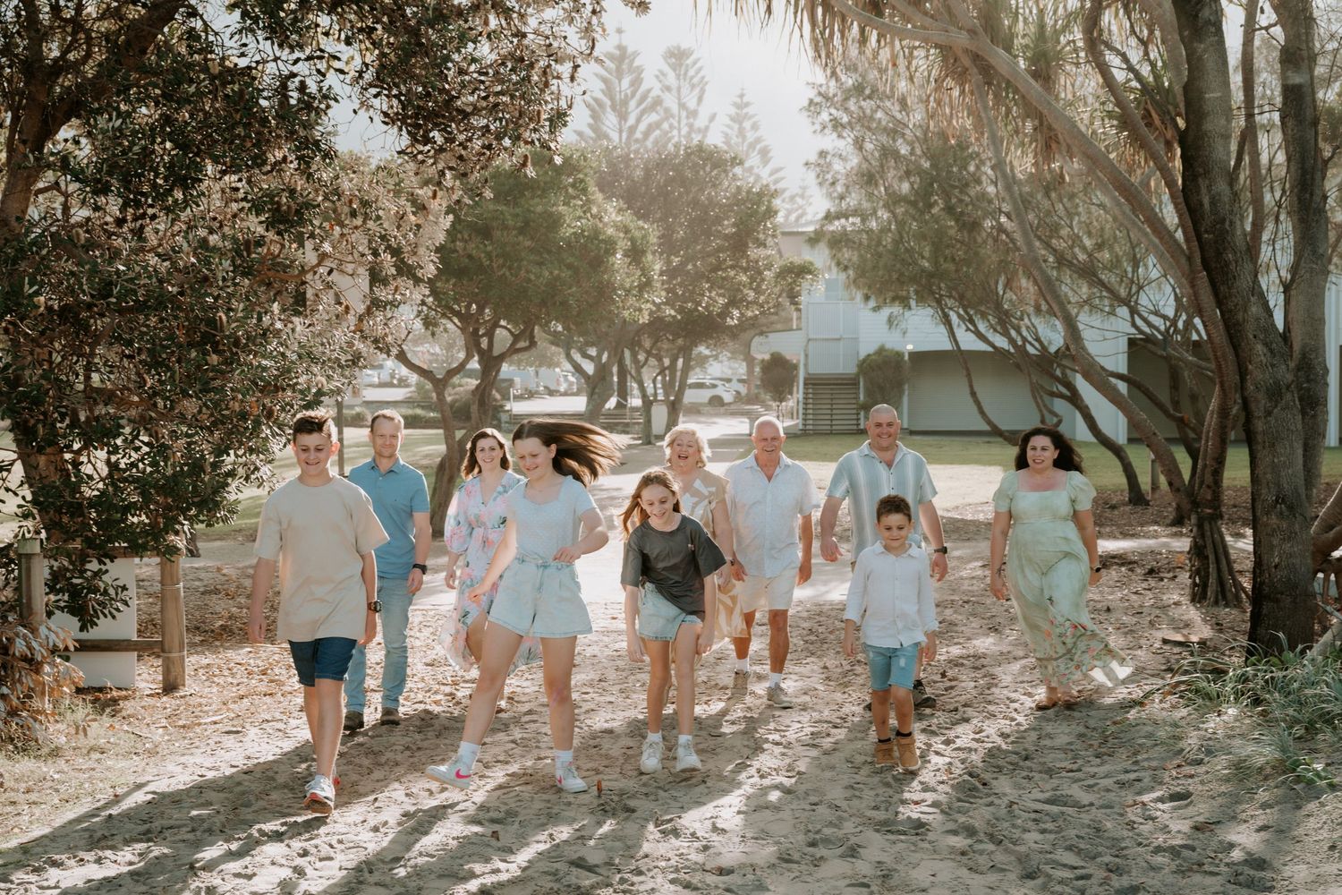 A large multigenerational family walks together on a sandy path surrounded by trees in a bright outdoor setting.