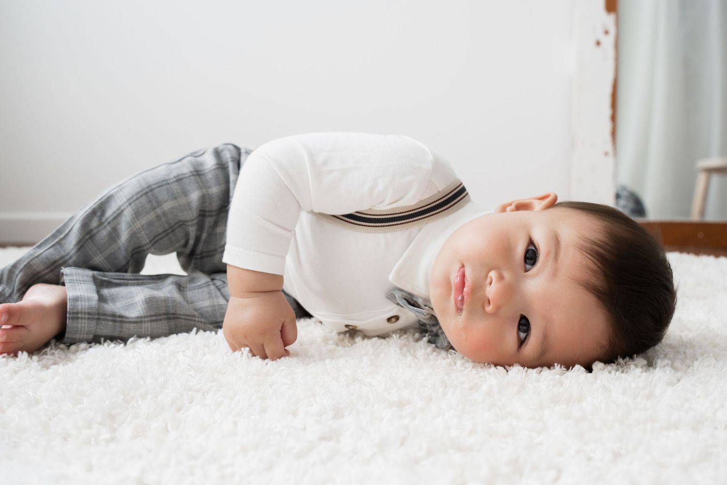 A cute baby in a white onesie and plaid pants lying on a soft white carpet, looking up with big brown eyes.