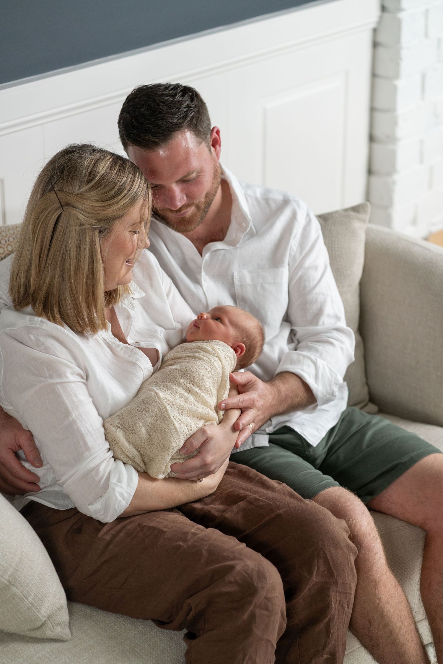 A couple sits on a couch, lovingly gazing at their newborn baby wrapped in a cream swaddle.