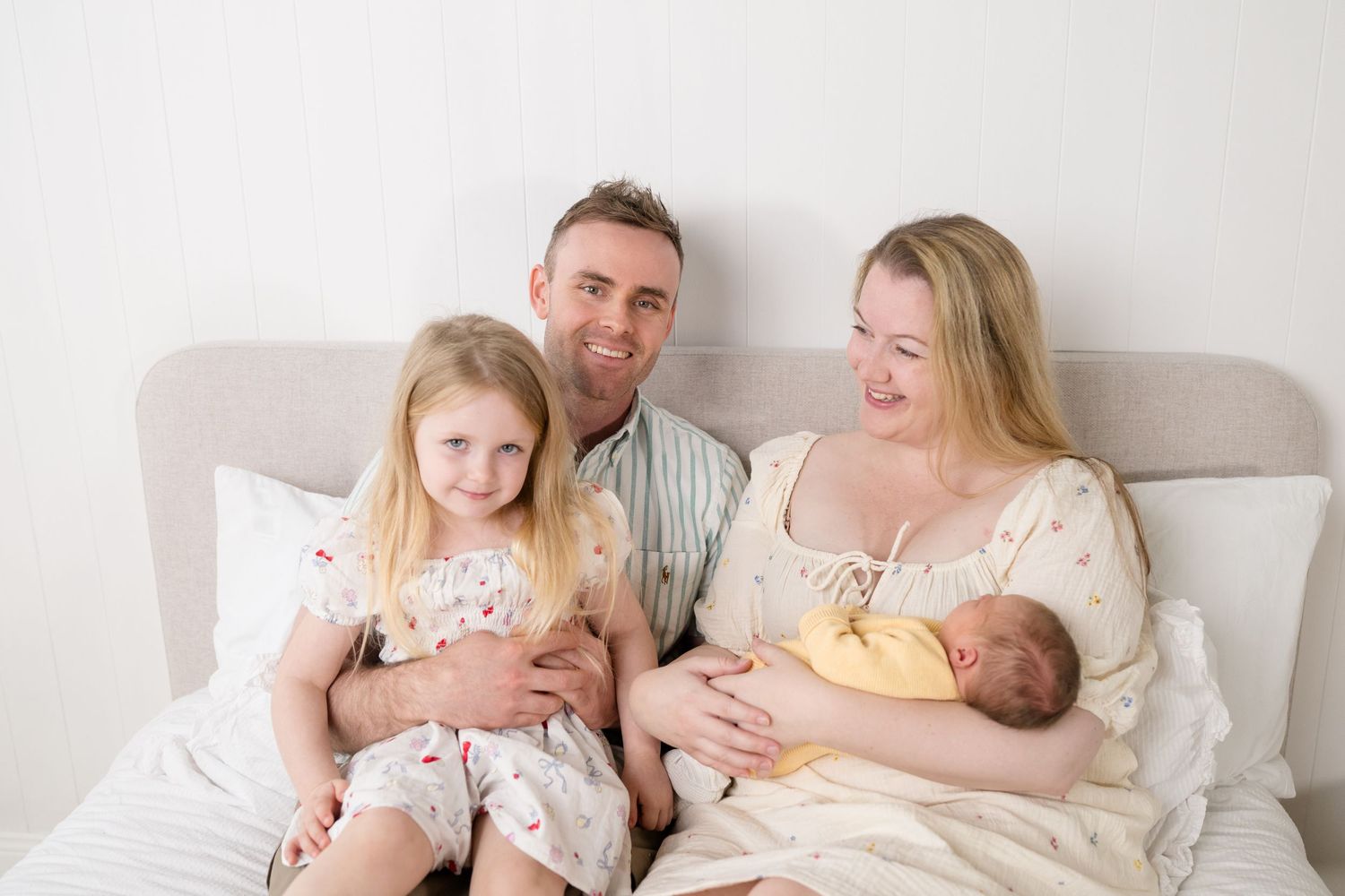 Happy family of four sitting on a white bed, parents smiling with young daughter and newborn baby in bright studio setting.