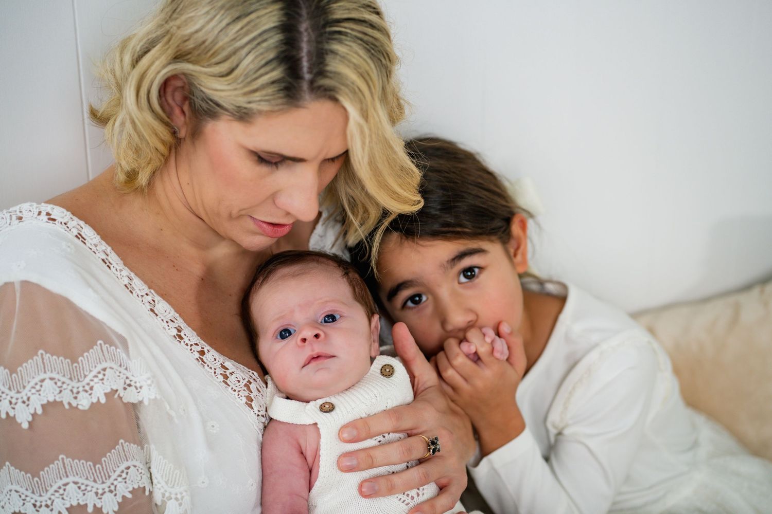 A mother in white holds her newborn baby while an older child leans in close, sharing a tender family moment.
