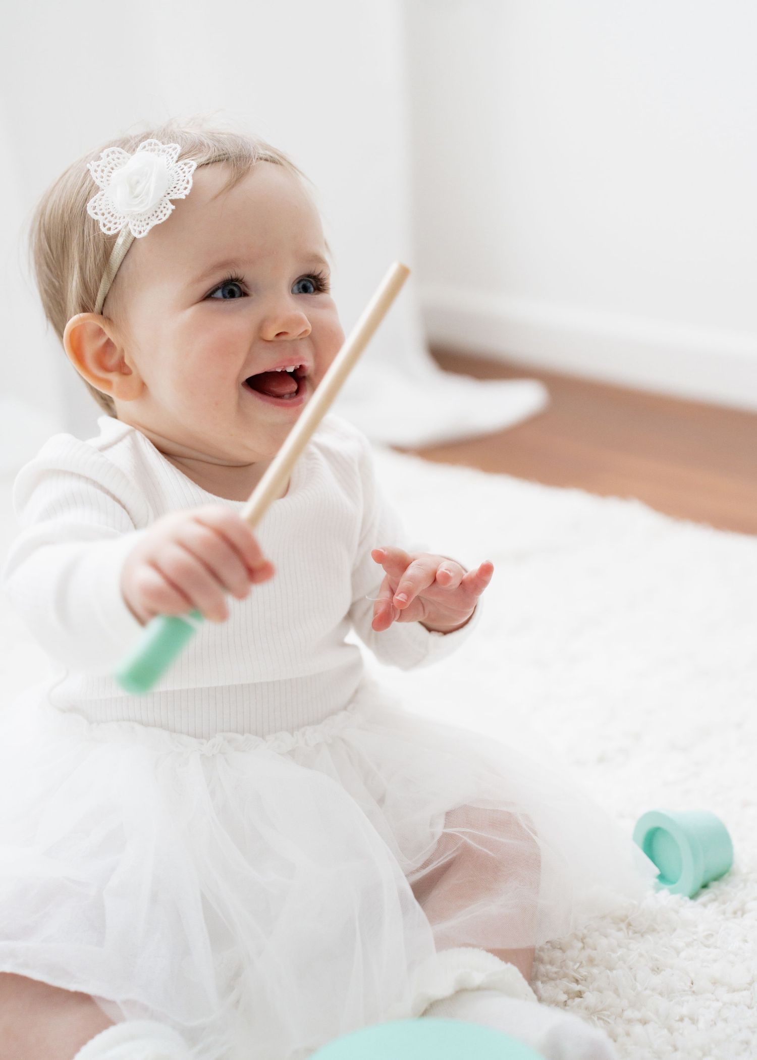 Happy baby girl in white dress and flower headband playing with wooden stick toy while sitting on fluffy rug.
