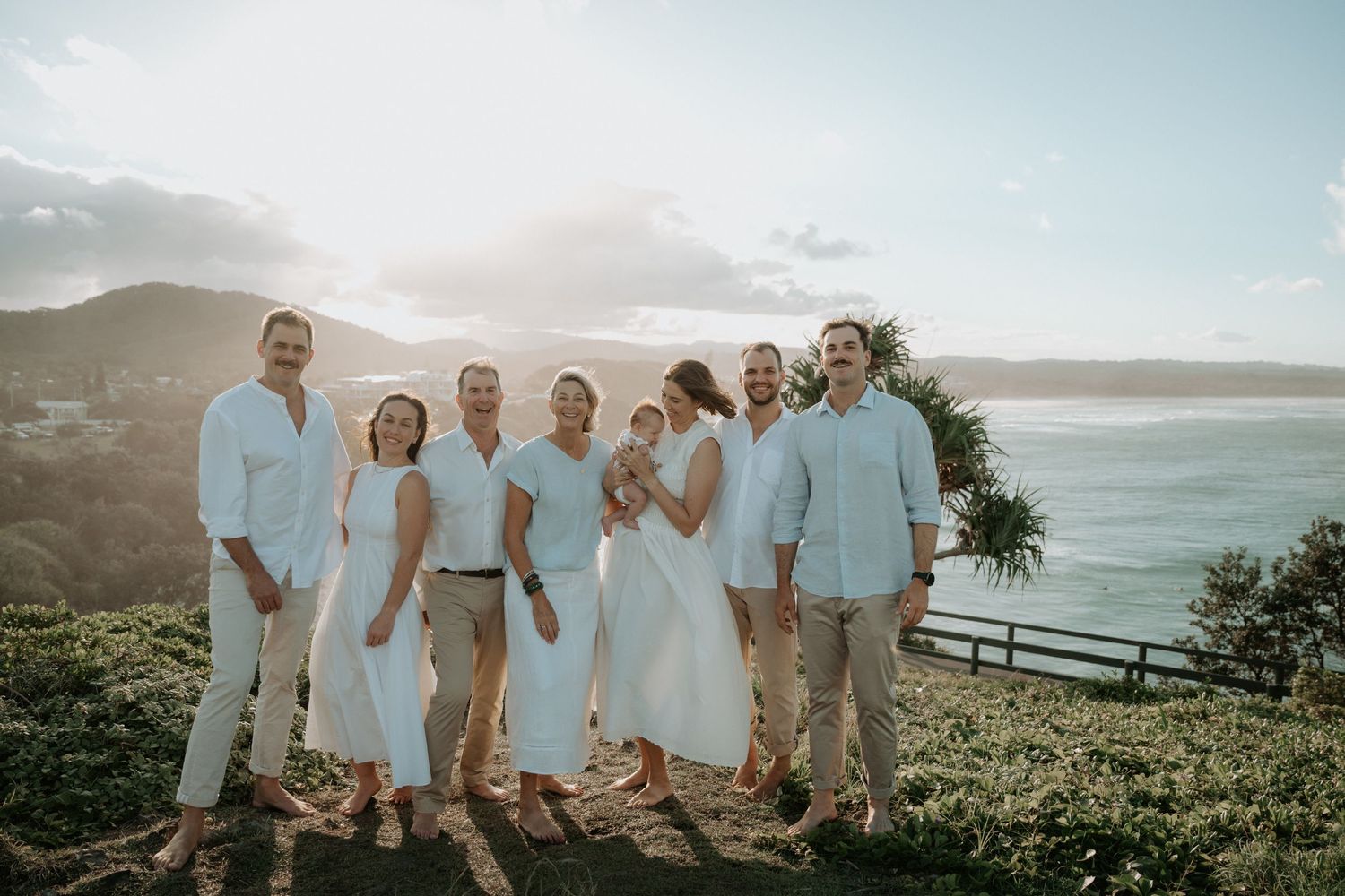 Family group photo outdoors at a coastal overlook, wearing white and beige outfits with ocean and mountains in background.