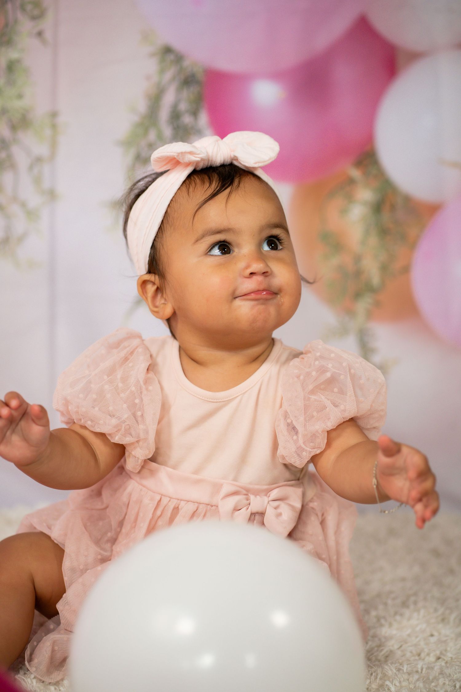 Adorable baby girl in pink lace dress and bow headband sitting among pink and white balloons.