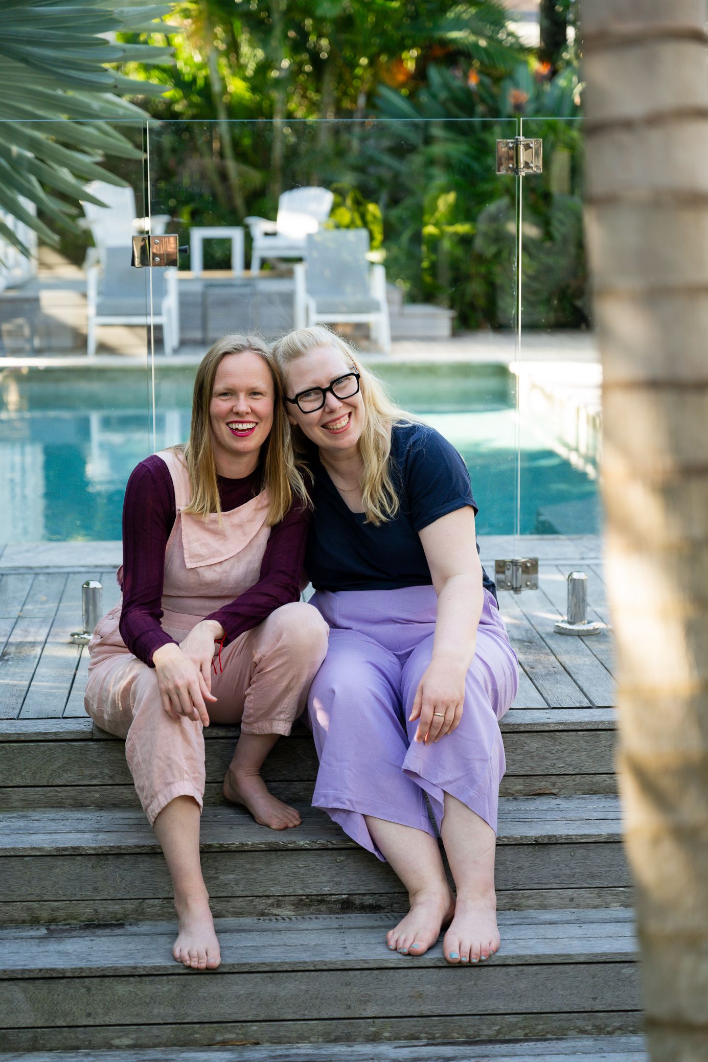 Two people in casual attire sit together barefoot on wooden deck steps by a pool on a sunny day.