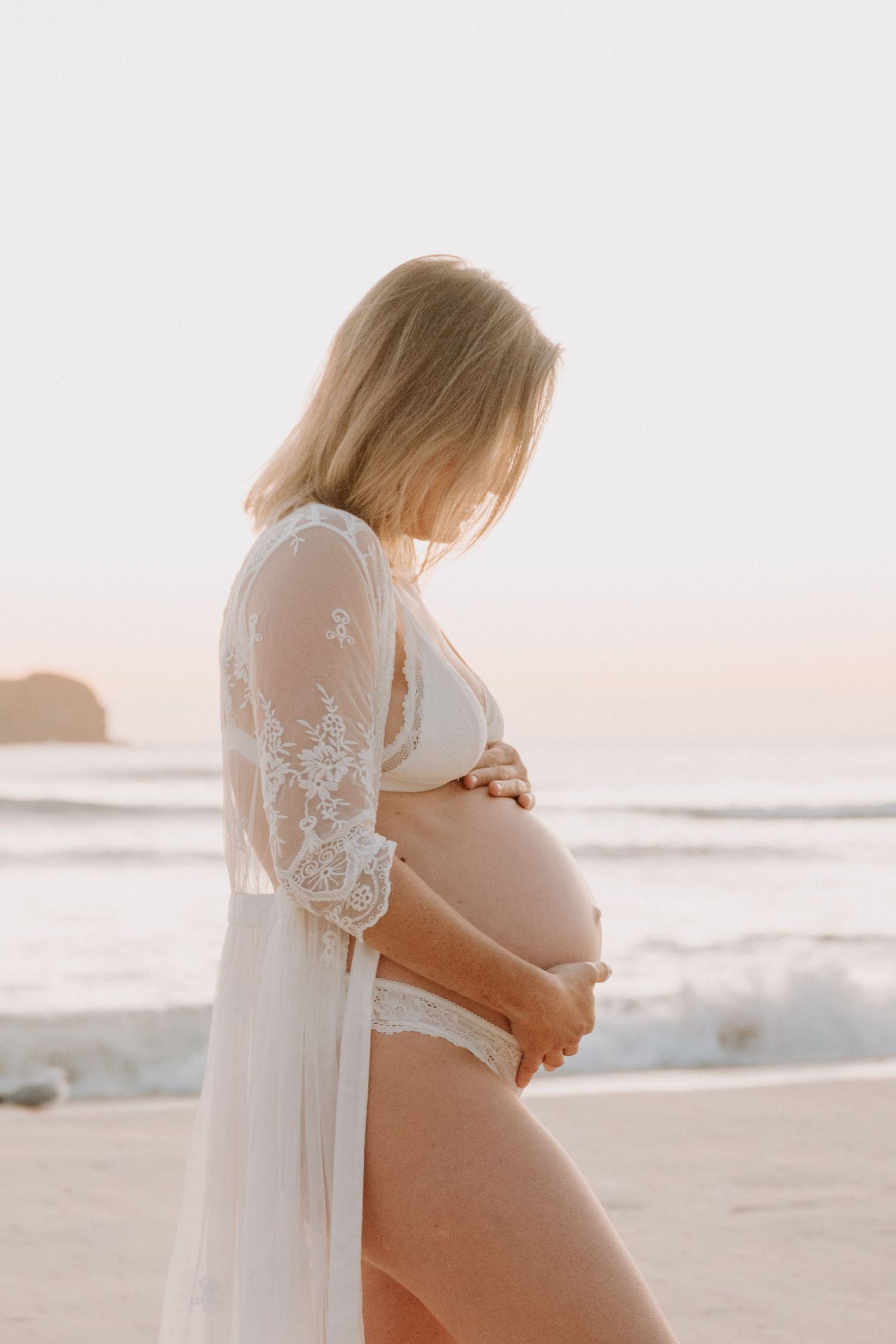 Pregnant woman in white lace robe and bikini cradling her baby bump on a serene beach at sunset.
