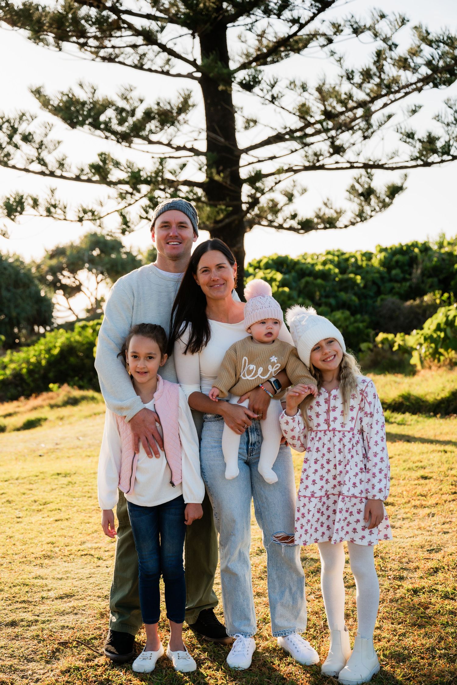 Family of five posing outdoors near a large pine tree on a sunny day, smiling together on green grass.