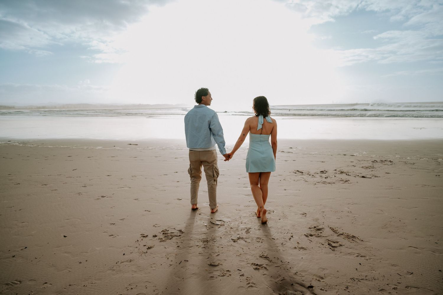 Couple holding hands walking toward ocean on sandy beach, backs to camera, romantic coastal scenery.