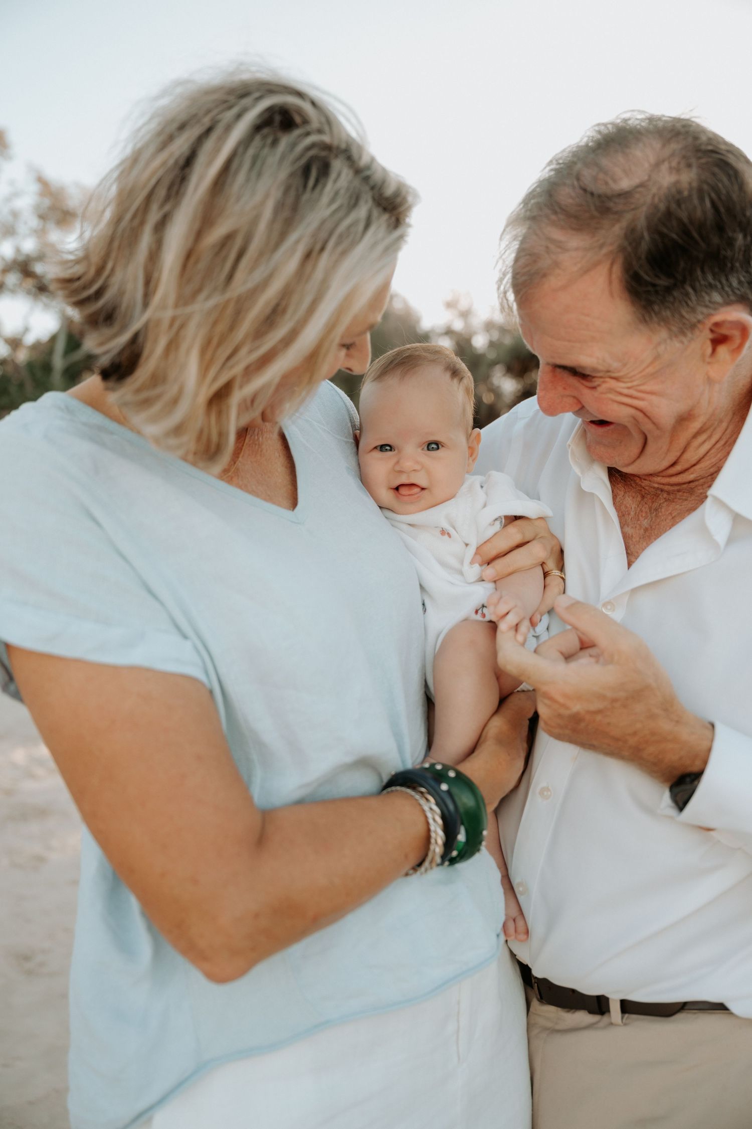 Grandparents smiling and holding a baby outdoors on a sunny day at the beach.