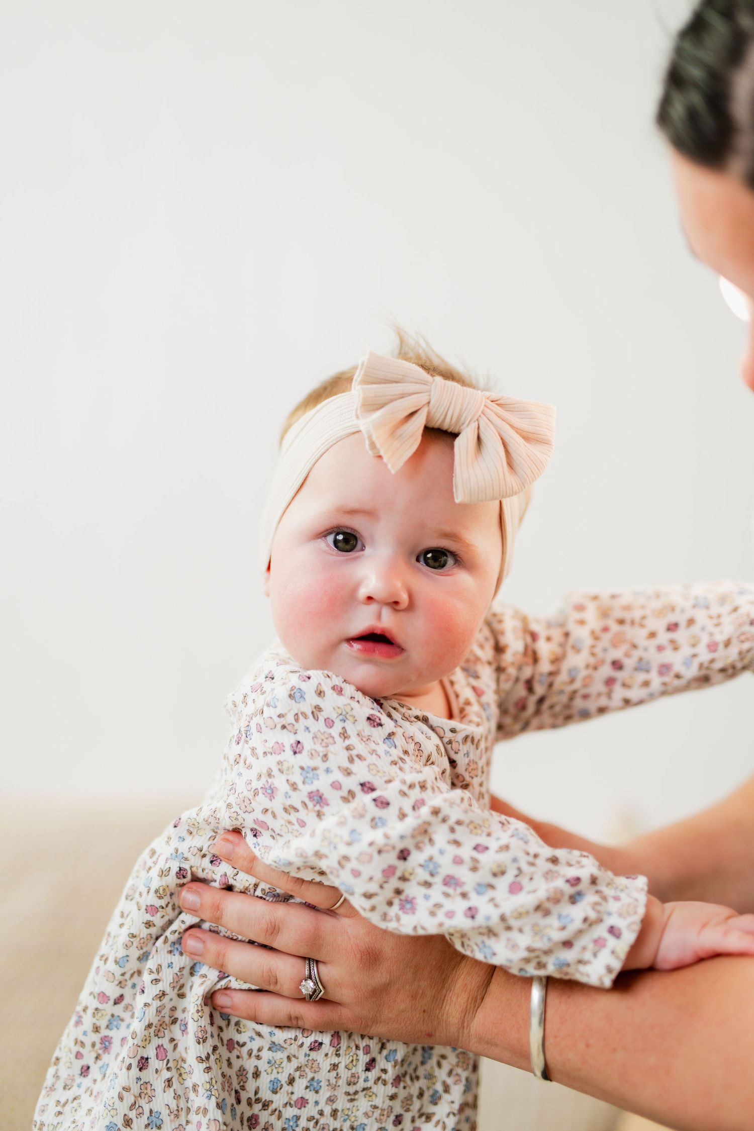 Adorable baby girl in floral dress and pink bow headband being held by mother against white background.