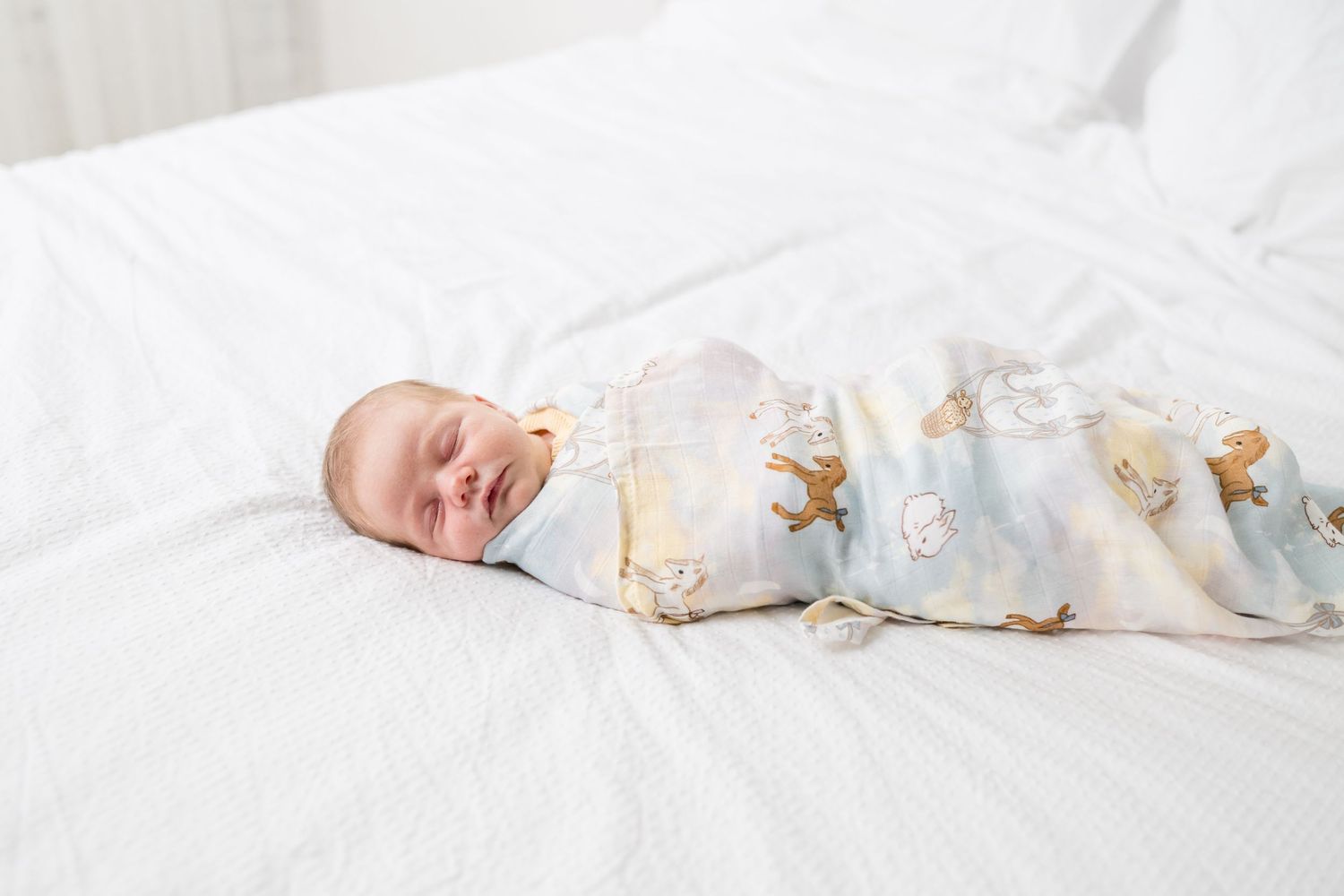 Newborn baby swaddled in a pastel animal print muslin blanket sleeping peacefully on a white bed.