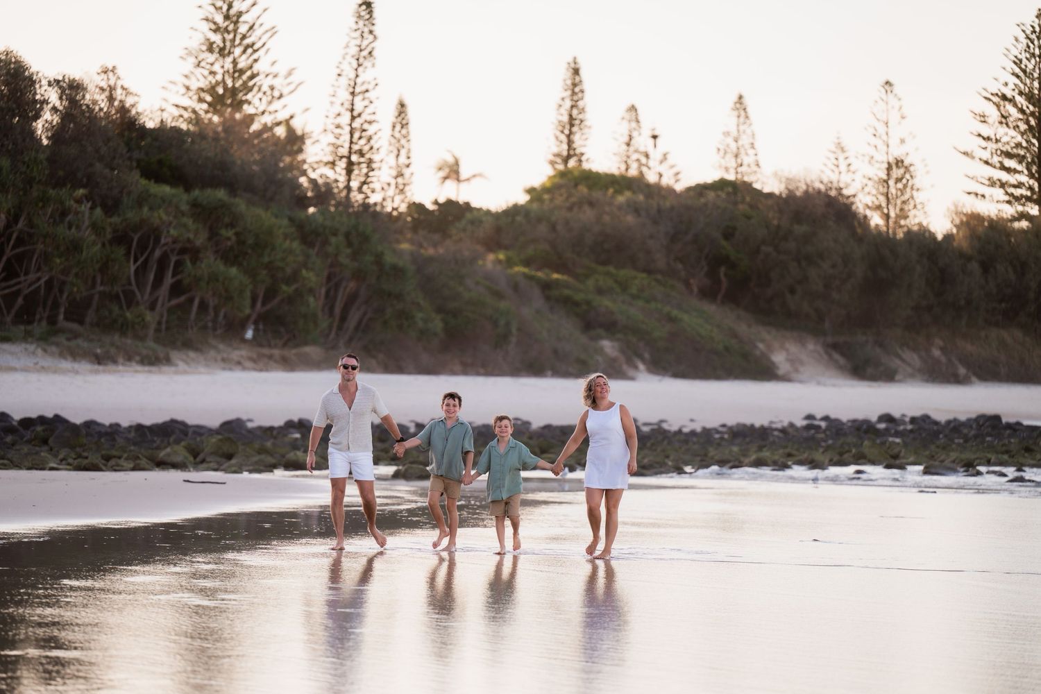 Family walking together on beach with pine trees in background during sunset.