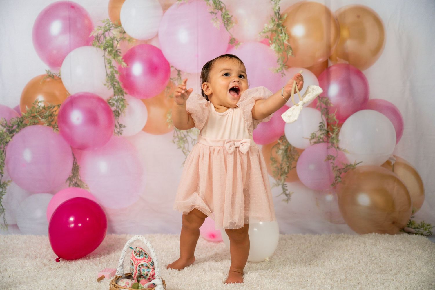 A baby girl in a pink dress celebrates her first birthday surrounded by pink, white, and gold balloons.