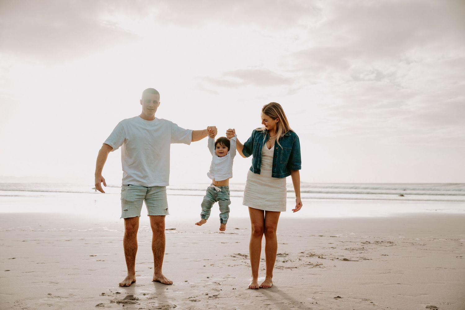 Family swinging toddler on sandy beach, parents holding child's hands with ocean waves in background.