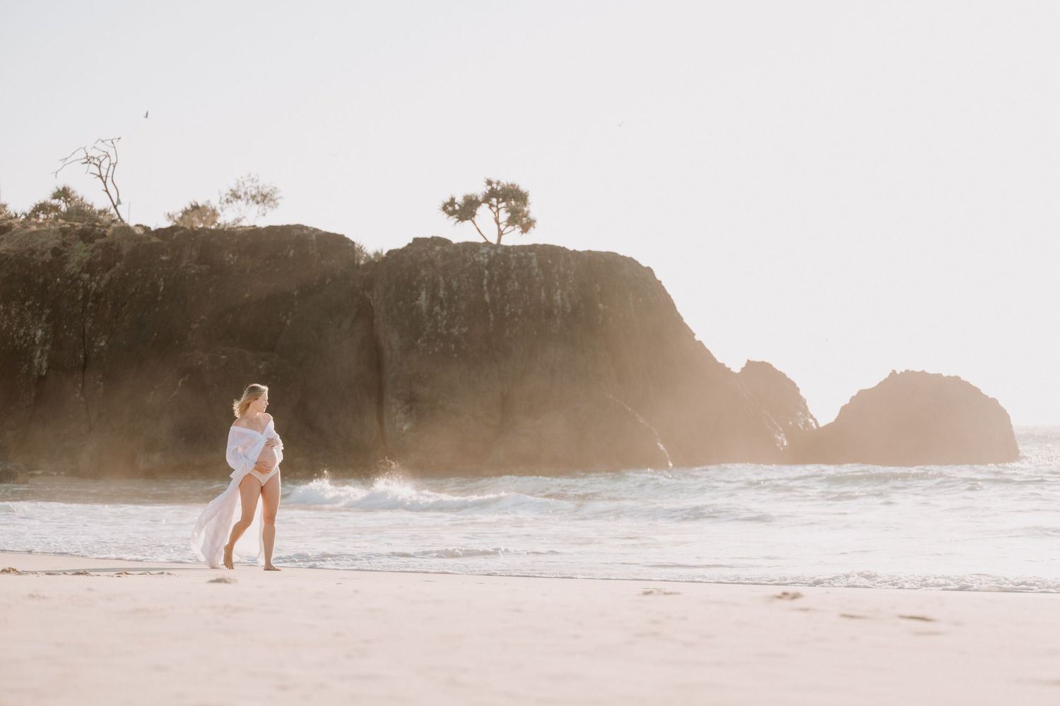 A woman walks along a sandy beach near rocky cliffs and ocean waves in warm golden sunlight.