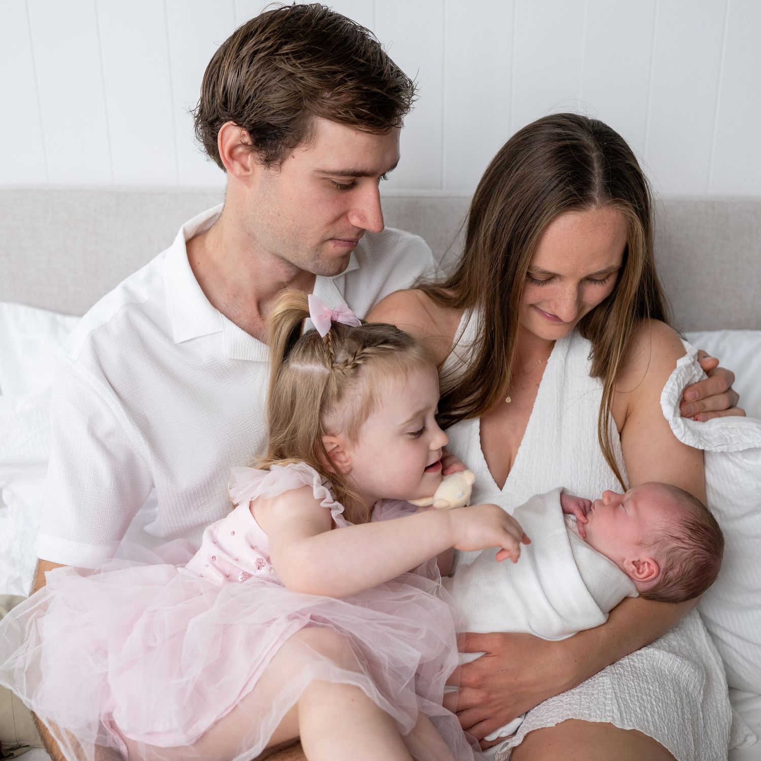 A family of four poses on a bed, parents in white holding a newborn while their toddler daughter in pink tutu looks on.
