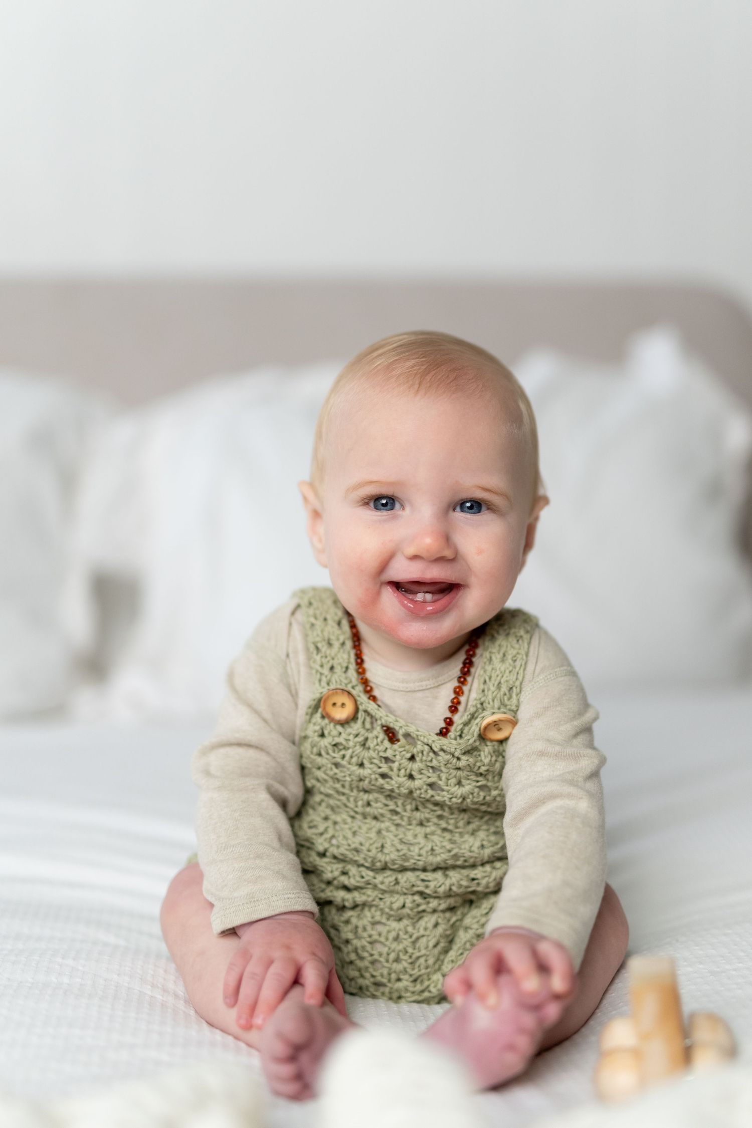 Smiling baby in green crochet overalls sitting on white bed.