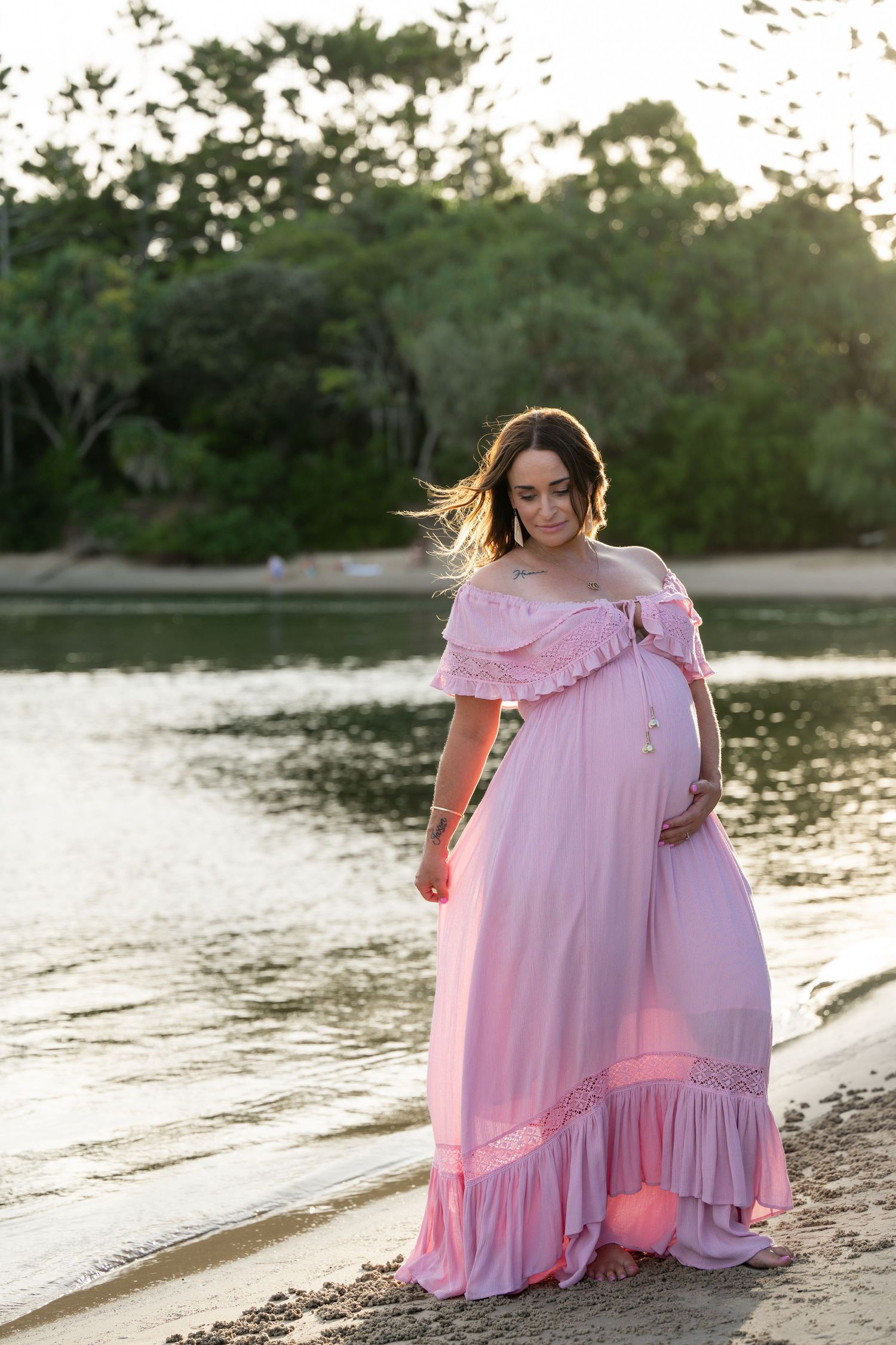 Pregnant woman in pink off-shoulder gown standing by a serene lake at sunset, cradling her baby bump.