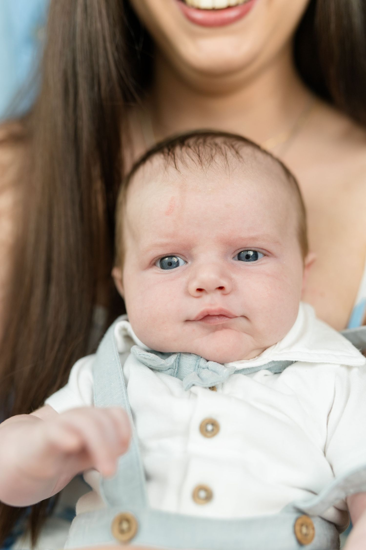 A smiling mother holds her newborn baby boy dressed in a white shirt and blue suspenders, looking at the camera.