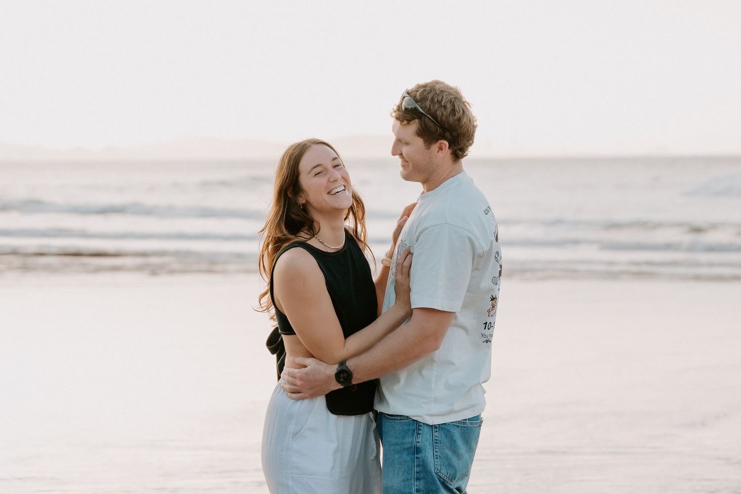 A happy couple embraces and laughs together on a sandy beach at sunset, with ocean waves in the background.