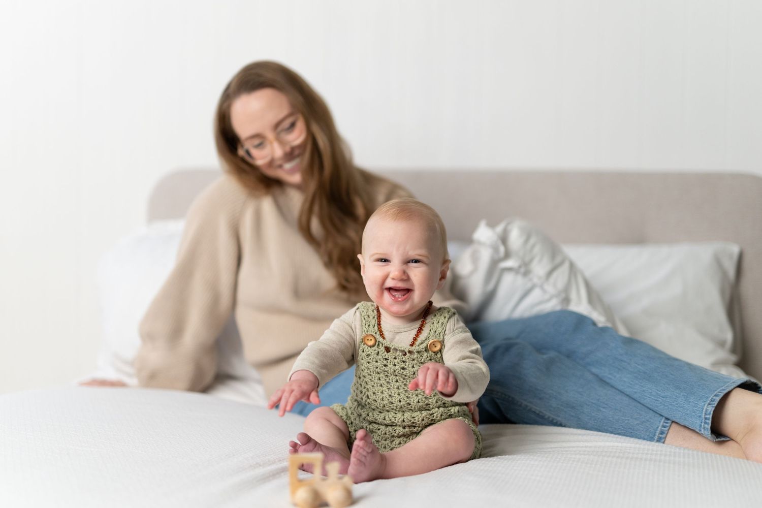 Happy baby sitting on white bed laughing while mother relaxes in background in bright minimalist room.