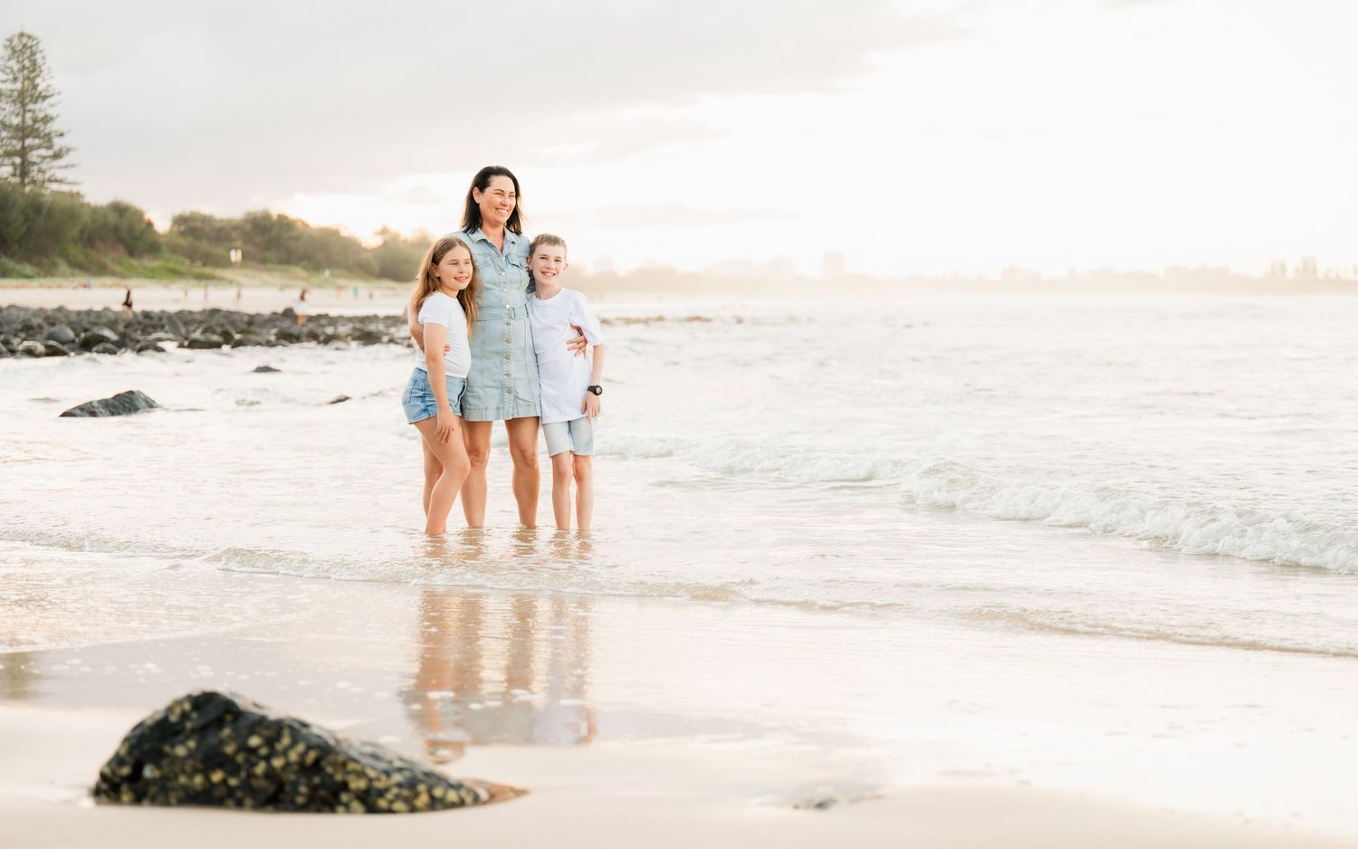A mother and two daughters stand together smiling on a sunny beach, wearing matching light blue outfits.