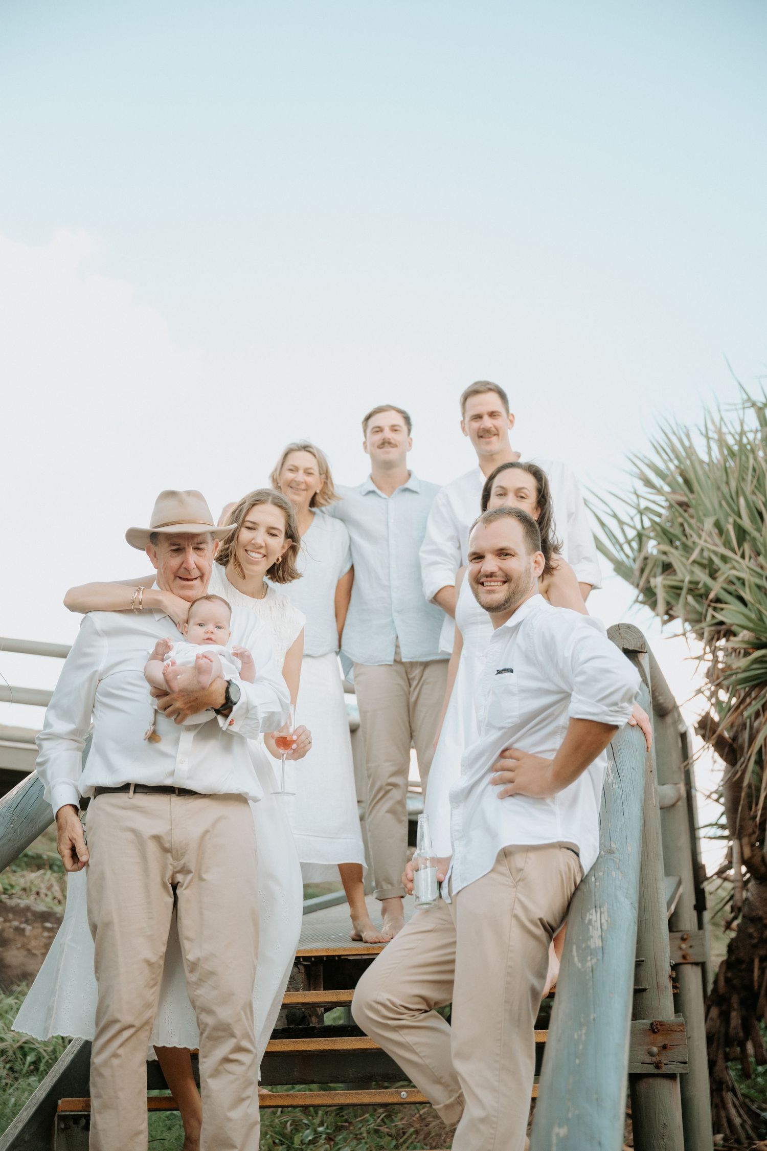 Family group photo on outdoor stairs in white and beige outfits, holding a baby, with tropical greenery background.