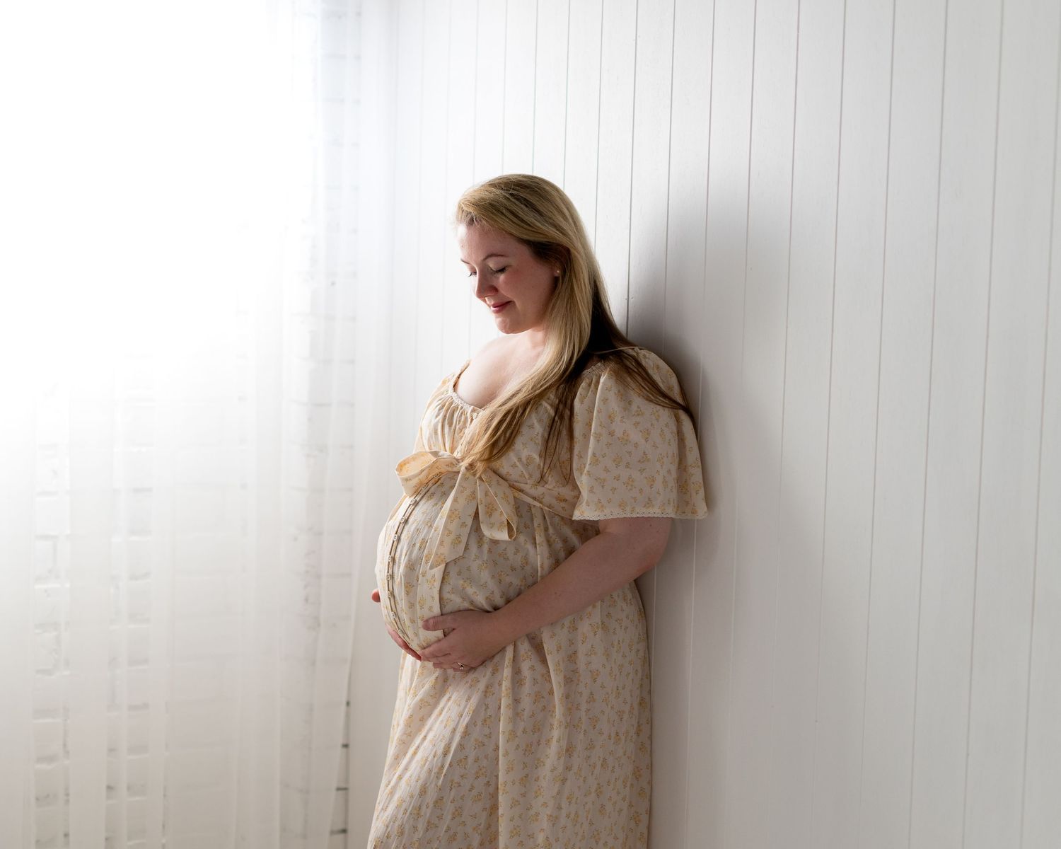 Pregnant woman in floral dress cradling baby bump, leaning against white wall near sheer curtain.
